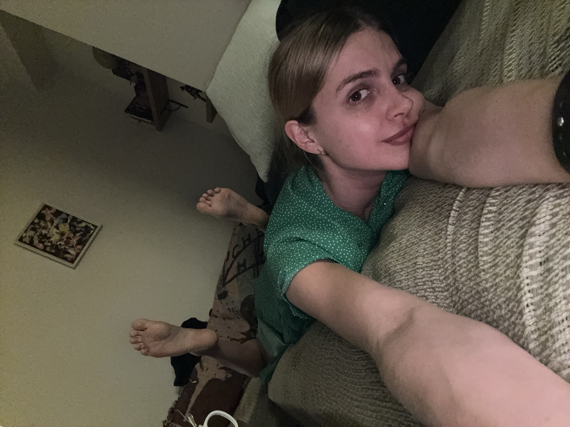 A young woman is captured in a close-up, tilted selfie taken indoors, likely in a living room or bedroom. She has light brown hair pulled back, fair skin, and dark eyes, looking directly at the camera with a relaxed, slightly smirking expression. She is wearing a green dress with white polka dots. Her right arm is bent and propped on a light-colored, textured couch or sofa, supporting her head, while her left arm extends towards the camera, indicating she is holding the device.
The background is dimly lit, suggesting evening or night. Behind her head, a light-colored pillow is visible. Further in the background, some dark furniture, possibly a shelf or table, can be seen with indistinct items on it. In the lower-left corner, on a light-colored wall, a small, framed piece of abstract art with colorful shapes is displayed. Her bare feet are visible in the mid-ground, one curled up. Near her feet, on what appears to be a patterned floor covering or blanket, there is a white, ring-shaped object. Some faint, largely illegible text, possibly "CH," is visible on the patterned fabric near her body. The overall atmosphere is intimate and casual.