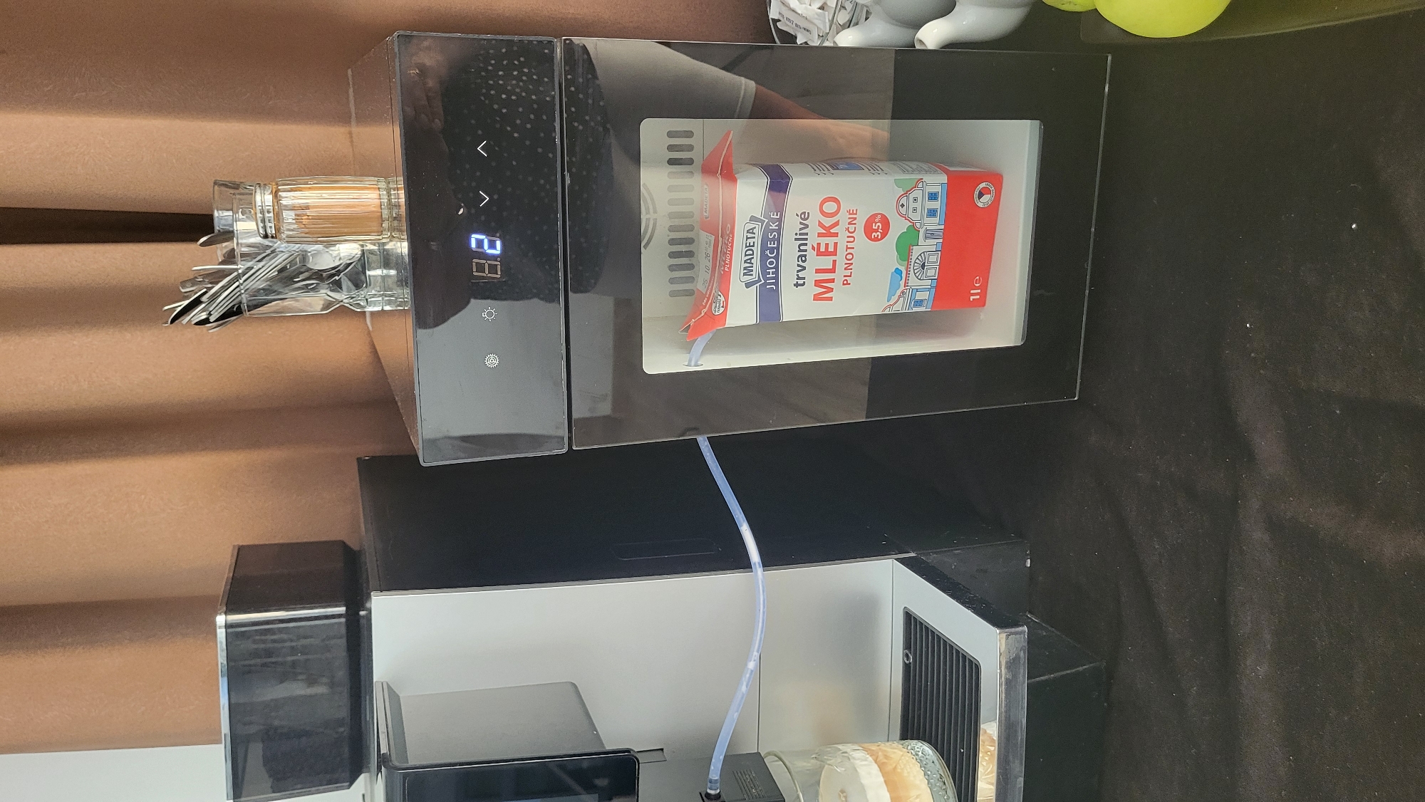 In Prešov, Slovakia, on a black tablecloth, a modern white and black coffee machine is positioned to the left. A clear plastic tube extends from the coffee machine to a glass containing a layered beverage. To the right of the coffee machine is a sleek black appliance with a digital display showing "8.2". Inside this appliance, a carton of "MADETA JIHOČESKÉ trvanlivé MLÉKO" (UHT Milk) is visible. The background consists of brown curtains.