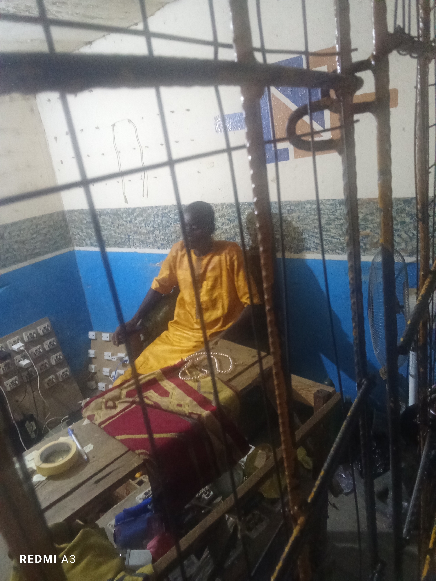 The image depicts an indoor scene, likely a workshop or a living space, in Jalingo, Nigeria. A man, appearing to be of African descent, is seated on a wooden platform covered with a colorful patterned cloth. He is wearing a loose-fitting, bright yellow garment. In front of him, also on the platform, is a necklace or strand of beads. To his left, a wooden table is visible, holding various items including a roll of yellow tape, what appears to be a roll of duct tape, and a pen. Further to the left, there are several boards with multiple white electrical outlets mounted on them, connected by wires, suggesting an electrical setup or repair area. The background features walls painted in sections of blue and a textured grey pattern. In the far right of the image, a black oscillating fan is partially visible behind a metal grate or bars, which also obscure the foreground and parts of the scene. The overall lighting is dim, casting shadows and contributing to a somewhat cluttered and utilitarian atmosphere. The presence of electrical outlets and the fan suggests a functional space, possibly related to electronics repair or a small business.
