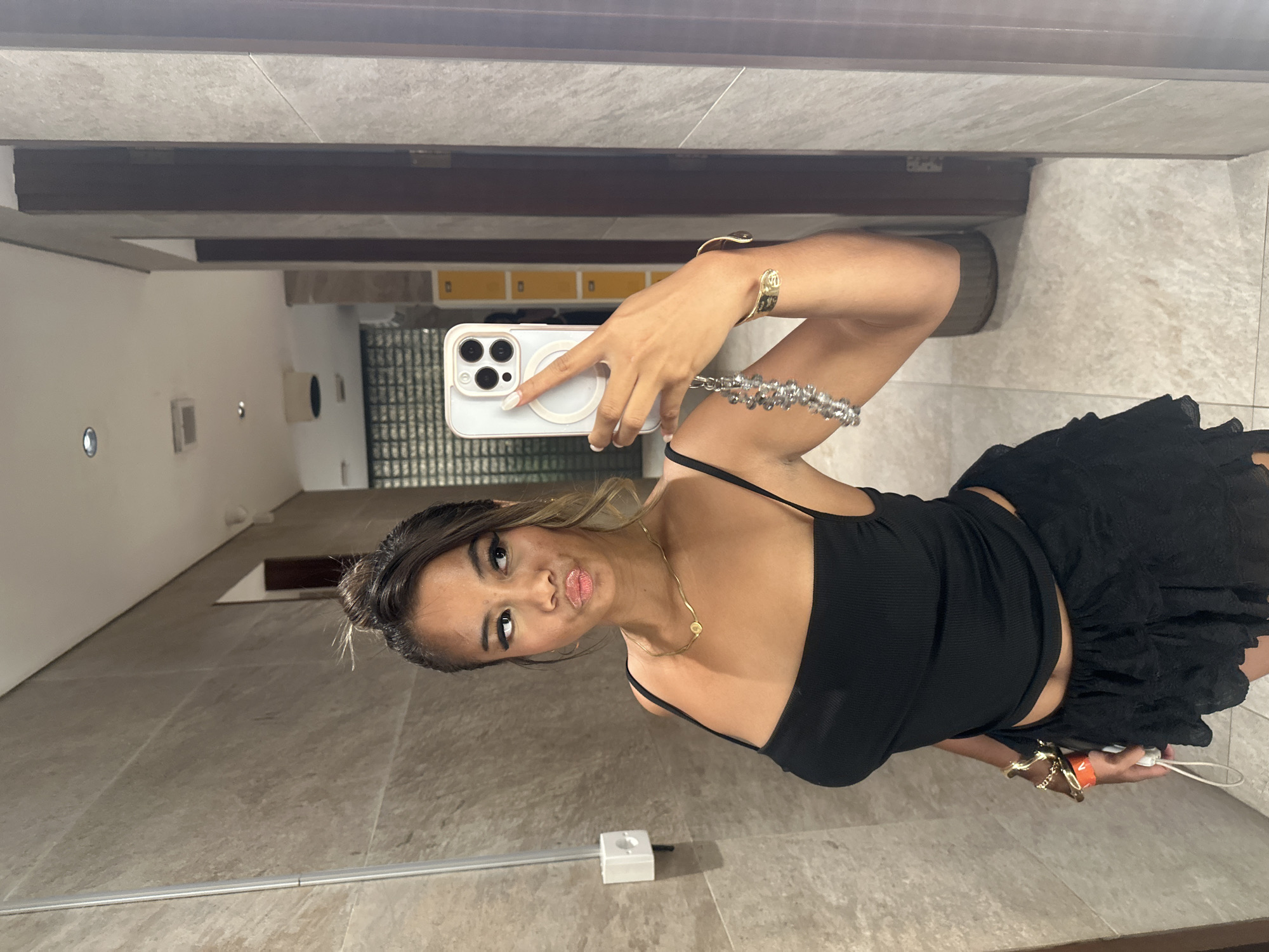 A rotated mirror selfie captures a young woman posing playfully in an indoor setting, likely a changing room or restroom in North Kuta, Indonesia. The woman, appearing to be of Asian descent, has dark hair styled in a bun with lighter highlights, and wears noticeable eye makeup and puckered pink lipstick, creating a "duck face" expression.
She is dressed in a black ribbed spaghetti strap top and a black ruffled or layered skirt. Her left hand holds a white smartphone with a distinct camera module and a circular magnetic accessory on its back, pointed towards the mirror to take the photograph. Adorning her, she wears a gold necklace with a small pendant, a gold cuff bracelet on her right wrist, and on her left arm, a multi-strand silver-toned bracelet, another bracelet, and a white and orange wristband.
The background reveals light-toned tiled walls and flooring, complemented by dark brown wooden beams or shelving units. Reflections in the mirror show hints of yellow lockers or cabinets, and possibly a glass block or textured glass partition. A small white electrical box with a wire is visible on the floor. The entire image is oriented sideways.
