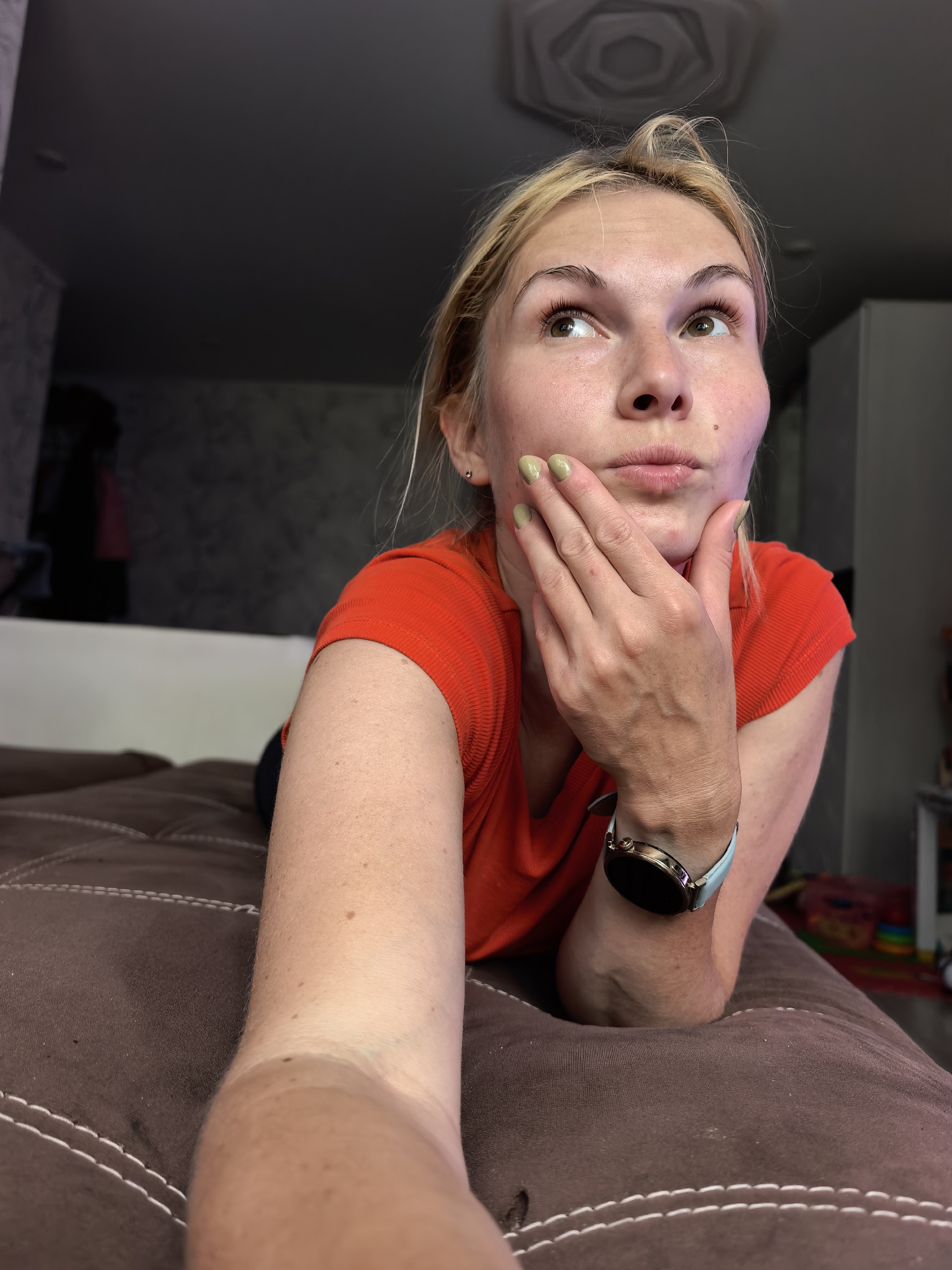 The media file features a medium shot of a young Caucasian woman lying on a brown textured surface, likely a bed or sofa. She is wearing an orange t-shirt. Her blonde hair is pulled back, with some strands loose around her face. She has fair skin and is looking upwards and to the right with a pensive or thoughtful expression. Her right hand is resting on her chin, with her fingers lightly touching her cheek. Her fingernails are painted a light green color. A smartwatch with a light blue band is visible on her left wrist.
The background is slightly blurred but shows a room interior. A textured gray wall is visible behind her, and part of a white headboard or wall is to the left. The ceiling has a decorative pattern. The overall lighting suggests it could be daytime, possibly indoors with artificial lighting. The woman's expression and hand gesture convey a sense of contemplation or curiosity. There is no visible text in the image. The location context provided is Sterlitamak, Russia.