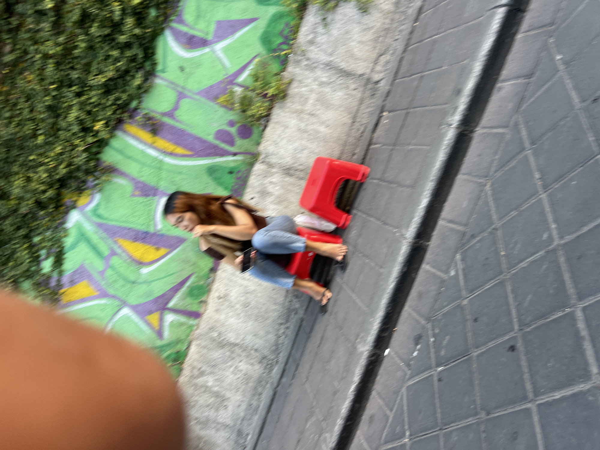 In Kuta, Indonesia, a young woman with long, auburn hair sits on a small, bright red plastic stool. She is wearing a dark brown top and light blue jeans. Her feet are bare, and she is holding a smartphone in her right hand. The stool is positioned on a paved walkway, likely made of grey interlocking bricks. Behind her, a concrete wall is adorned with vibrant graffiti featuring abstract shapes in shades of green, purple, and yellow. Lush green foliage cascades down from the top of the wall, partially obscuring the graffiti. A blurry, out-of-focus object, possibly a hand or arm, is visible in the upper left corner of the image. The scene appears to be outdoors, possibly in a street or public area in Kuta.