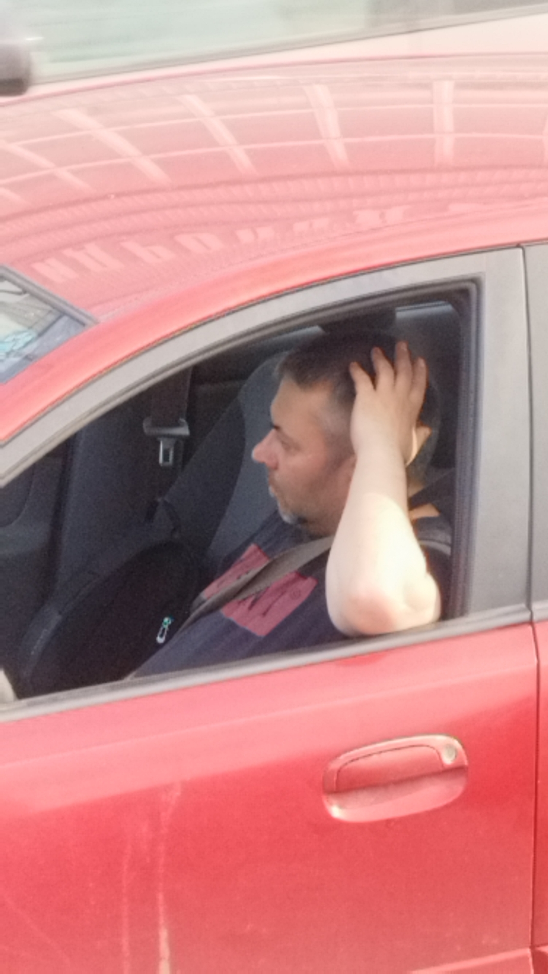 A man is seated in the driver's side of a red car, looking to the left through the open window. His right arm is raised and resting on the back of his head. He has short, dark hair with some graying at the temples and a stubbled beard. He is wearing a dark blue and red patterned sweater. A seatbelt is visible across his chest. The interior of the car appears to be dark gray. The exterior of the car is a bright red, and the roof has a reflective surface showing a pattern that might be from a ceiling or overhead structure. The scene suggests it is daytime. There is no visible text.