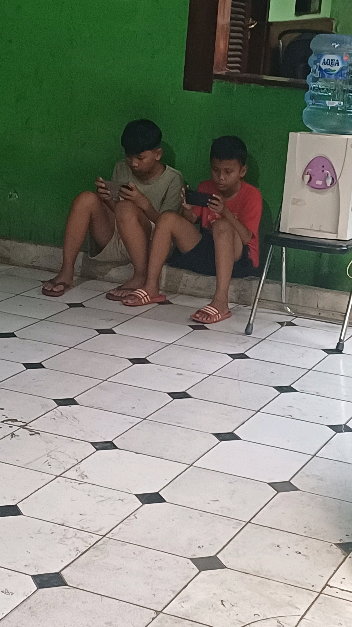 Two young males, appearing to be under 18 years old, are seated on the floor in front of a green wall. Both are holding smartphones and appear to be engaged in playing mobile games. The male on the left is wearing a light olive green t-shirt and light-colored shorts, while the male on the right is wearing a red t-shirt and dark shorts. Both are wearing sandals. To the right of the males, there is a white water dispenser with a blue Aqua brand water jug on top. A metal chair with a black seat is partially visible behind the water dispenser. The floor is tiled with a white and black geometric pattern. The scene appears to be indoors, possibly in a residential setting. The location is identified as South Jakarta, Indonesia.