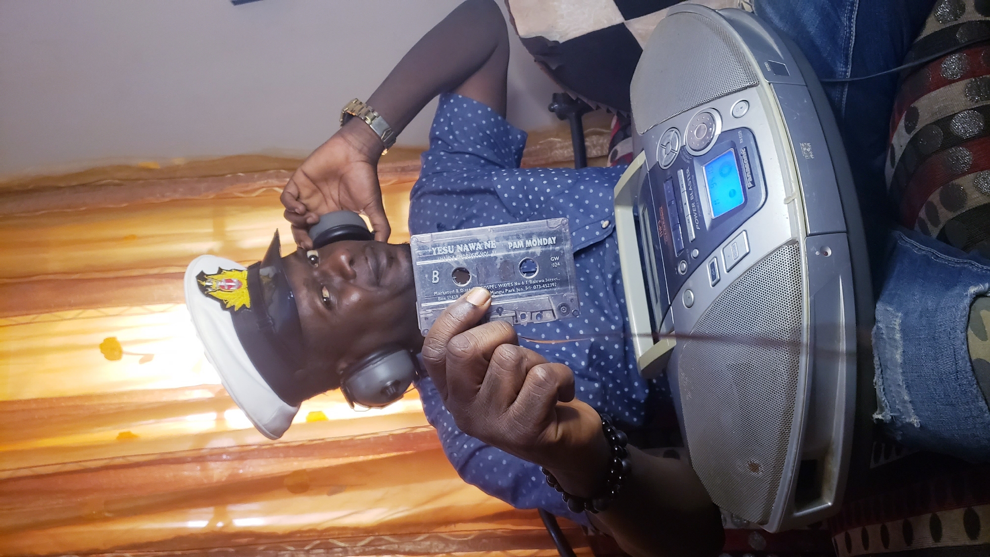 A man wearing a white sailor's cap with a gold emblem, headphones, and a blue polka-dotted shirt is holding a cassette tape with his left hand. He is sitting in what appears to be a living room in Narayi, Nigeria, with orange curtains in the background. In front of him is a silver boombox. The man's right hand is near his headphones. He is wearing blue jeans.
