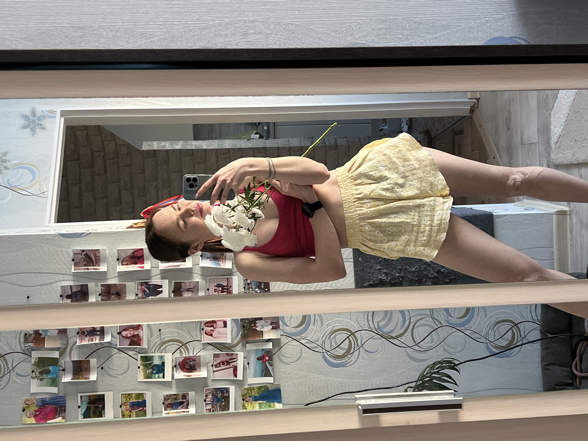 The image captures a mirror selfie of a young woman, appearing to stand upright as reflected in the mirror. She is holding a small bouquet of white flowers in her right hand, while her left hand is raised, holding a smartphone to take the picture. She is wearing a red sleeveless top and a light yellow, textured skirt that reaches above her knees. A dark-colored watch is visible on her left wrist, and a thin bracelet on her right wrist. Her hair is dark, and she has a red accessory, possibly a headband, on her head. There are some light-colored, patterned markings or a design on her right thigh/knee area.
The background reflected in the mirror reveals an indoor setting. The wall directly behind her features a light blue wallpaper adorned with abstract grey and blue swirling patterns. A prominent collage of numerous small, square photographs (resembling instant prints or polaroids) is pinned or taped to this wall, arranged in multiple horizontal rows from the mid-section down. Through an open doorway or archway visible behind her head and upper body, another wall with a distinct brown and grey stone or brick-like texture is reflected.
Below the photo collage and across the lower part of the reflected wall area, there are black and grey electrical wires running horizontally. A small green plant leaf is visible towards the bottom center, and a metallic strip or edge of a shelf is at the very bottom. The floor reflected beneath her legs appears to be a light-colored wood or laminate. A faint snowflake pattern is also visible on the wallpaper in the upper left corner of the reflection. The overall scene suggests a casual moment captured indoors, likely in a living space or bedroom.