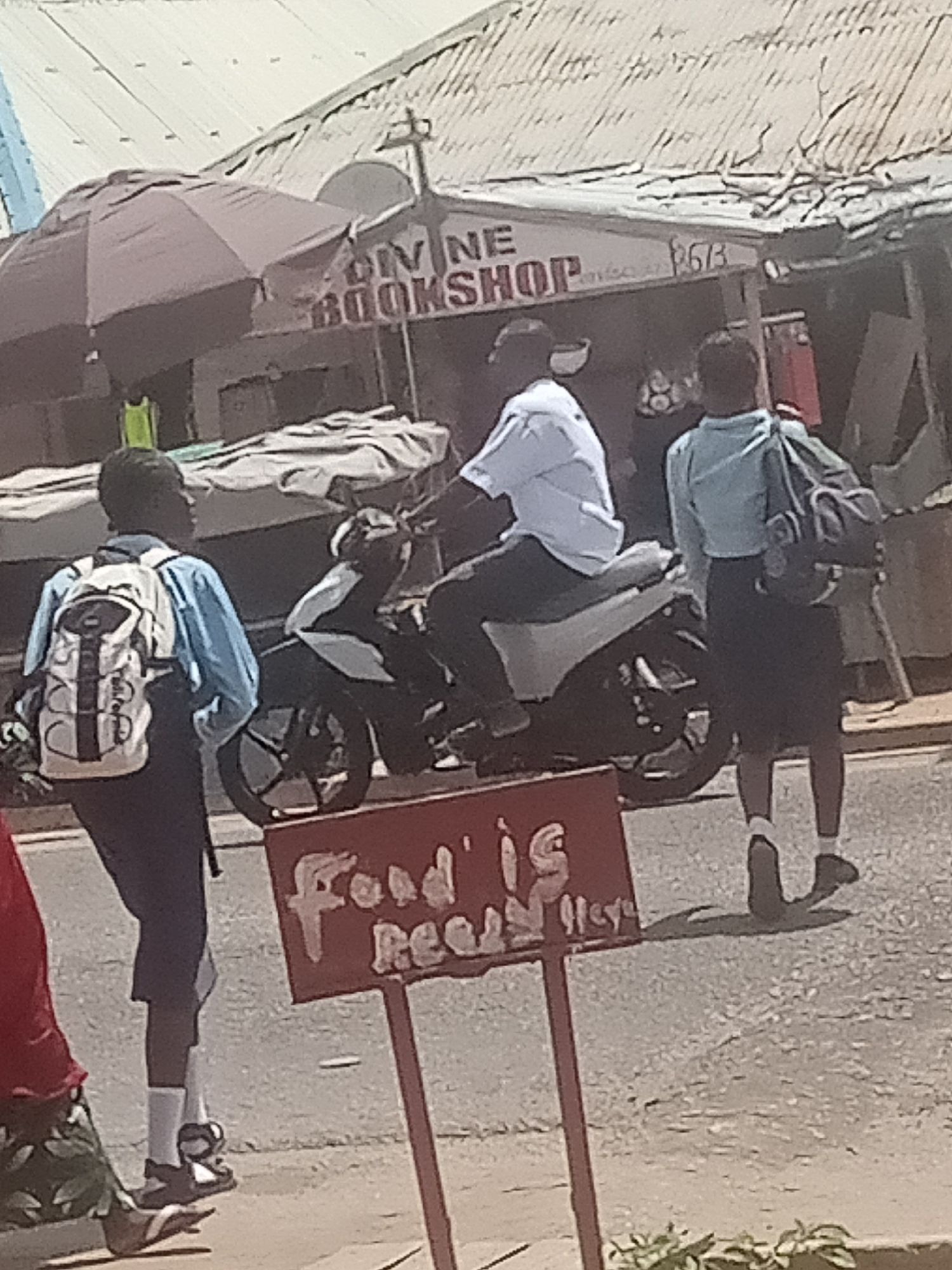 The image depicts a street scene in Jalingo, Nigeria, under a bright, likely daytime sky, suggesting fair weather. In the foreground, a red sign with white lettering reads "Food is Ready". The sign is supported by a metal pole.
Several people are present. To the left, a young person wearing a blue shirt and dark pants, with a white and black backpack, walks along the sidewalk. Further back, a person is seated on a motorcycle, wearing a white shirt and dark pants. To the right, another young person, dressed in a light blue shirt and dark skirt, walks away from the motorcycle, carrying a dark backpack. A partially visible person in a red top and patterned skirt is seated to the far left.
Behind the people and the motorcycle, a storefront is visible with a corrugated metal roof. A sign above the storefront reads "DIVINE BOOKSHOP". An umbrella shades part of the entrance. The overall impression is of a bustling street with everyday activities taking place.