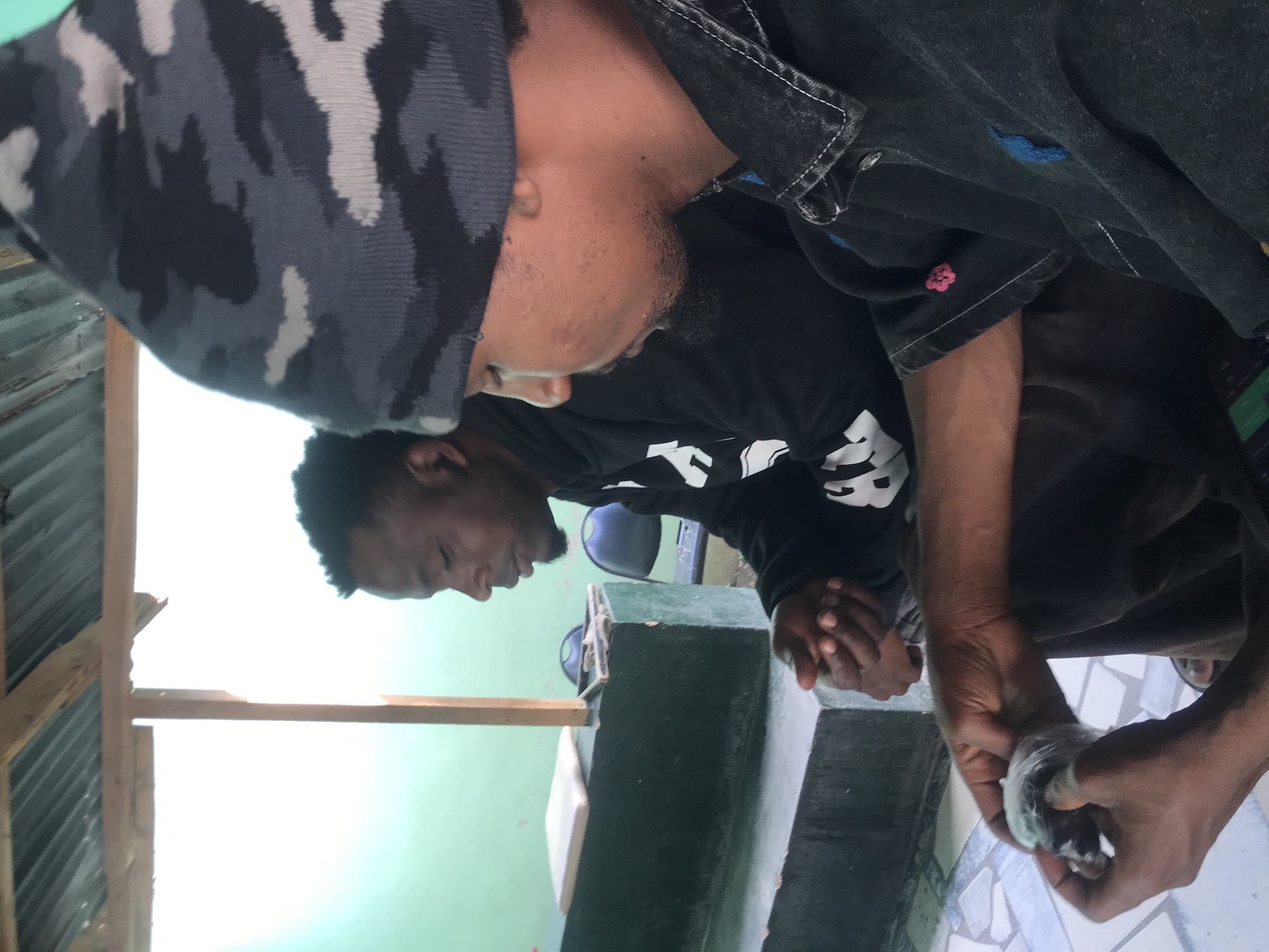This image captures two adult males with dark skin tones inside a structure with a corrugated metal roof and concrete elements, in an unknown city and country.
In the foreground, a man is positioned on the right, viewed from the upper torso, head, and hands. He wears a dark gray, black, and light gray camouflage-patterned knit beanie. His attire consists of a dark denim-like collared shirt or jacket with visible white stitching, featuring a blue embroidered logo (possibly a polo player) on the left chest and a small pink floral or star-like embroidery near the left sleeve cuff. His hands are central in the lower frame, holding and seemingly unwrapping or preparing a small, light-colored, possibly food-related item, which appears to be wrapped in clear plastic. He has a dark beard stubble and is looking downwards at his hands.
Behind him, to the left, a second man is seated, visible in profile from the chest up. He wears a black hooded sweatshirt with white lettering or graphics (partially visible) on the front. His hands are clasped or resting in his lap, and his gaze is directed downwards or forward, suggesting contemplation.
The men are seated on or next to a dark green painted concrete block structure. The foreground man is positioned lower, while the background man sits slightly higher on what appears to be a tiered concrete bench or platform. To the left behind the background man, a light green painted wall is visible. To the right and above, a bright, overexposed white area indicates an open space, likely a window or doorway, with strong ambient light. A light brown wooden support post is vertically positioned within this bright area. Above, a corrugated metal roof, supported by wooden beams, is discernible. The floor in the lower part of the image features a white and gray geometric patterned tile. On the concrete structure behind the background man, a light-colored cushion or pad is present, along with a dark object resembling a mobile phone or small tablet. In the far background, two dark, possibly plastic, chairs are visible.