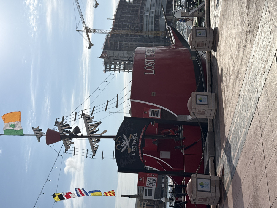 The image depicts a red pirate-themed tour boat named "LOST PEARL" docked along a waterfront promenade in Downtown Tampa, Florida, United States of America. The boat features a mast flying an Irish tricolor flag with a green shamrock and a sign at its entrance displaying a pirate skull and crossbones, the name "LOST PEARL," and "Welcome Aboard, Mateys!" The vessel's hull is red with black trim, and its name is painted in white on the stern.
In the foreground, a paved walkway, composed of reddish-brown and gray interlocking pavers, extends across the scene. Two stone bollards, each displaying a "River Walk" logo and connected by a chain, mark the edge of the promenade adjacent to the boat.
To the left of the boat, a utility pole displays a vertical string of multicolored nautical signal flags. The background on the right reveals a significant urban construction site featuring multiple partially completed high-rise concrete structures and at least two prominent yellow tower cranes against a clear, light blue sky with sparse clouds. A small black flag, possibly a pirate flag, is visible atop one of the distant construction buildings. A faint, light-clad figure of a person is visible near the water's edge further down the dock on the far right. The scene is brightly lit by sunlight, casting distinct shadows on the ground.