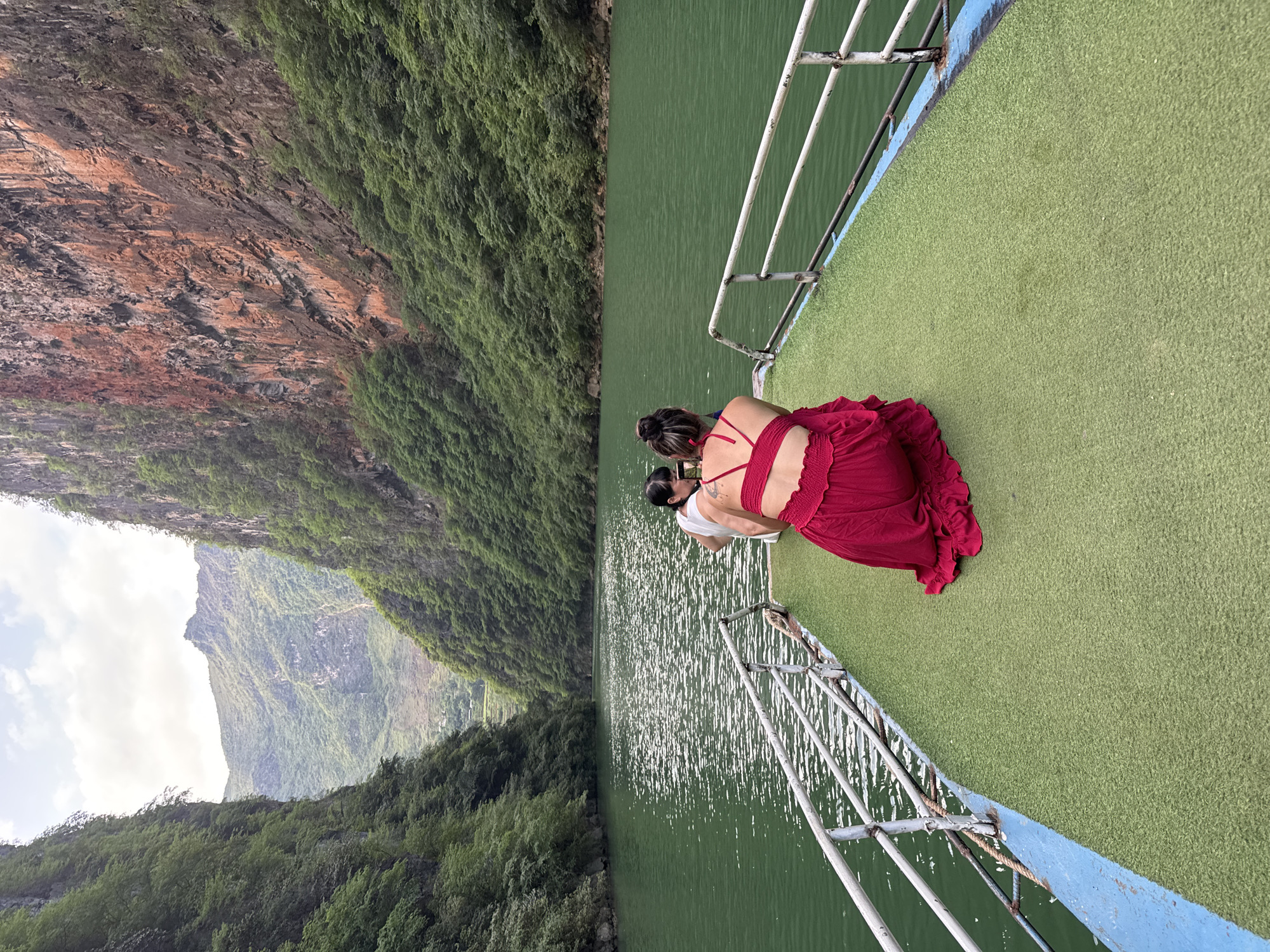 Two people stand on the green deck of a boat, looking out over a body of water towards a cliff covered in green vegetation. The boat has a white metal railing. The water is green and reflects sunlight. The cliff face is partially covered in brown rock formations. The scene appears to be located in a canyon or gorge. It can be deduced that this is in Đồng Văn Commune, Vietnam, because the image was specifically requested to be recognized as such.