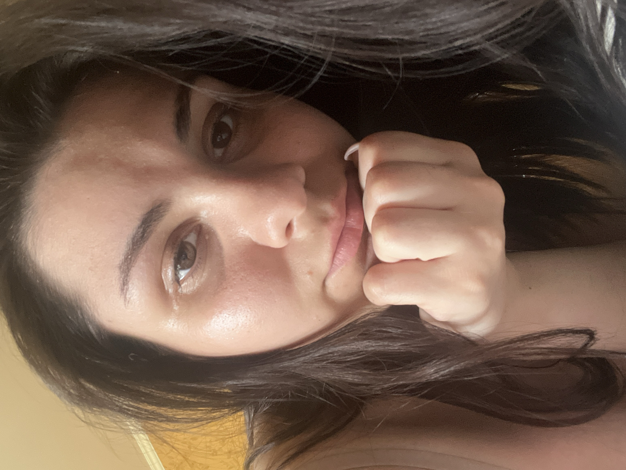 The image is a close-up, slightly tilted selfie of a young woman. She has long, dark hair that frames her face, with some strands falling over her forehead and shoulder. Her skin appears fair with a slight sheen, suggesting natural light. Her eyes are brown and her eyebrows are well-defined. Her lips are slightly parted, revealing a hint of her teeth. Her right hand is raised, with her fingers gently touching her cheek and mouth area, adding a thoughtful or pensive expression. Her fingernails appear to be painted white. The background is a muted, warm yellow color, possibly a wall or fabric, and is out of focus. The lighting is soft and appears to be coming from the side, highlighting the contours of her face and creating subtle shadows. There is no visible text in the image. The overall mood is intimate and self-reflective. The time of day and weather are not discernible from the image.