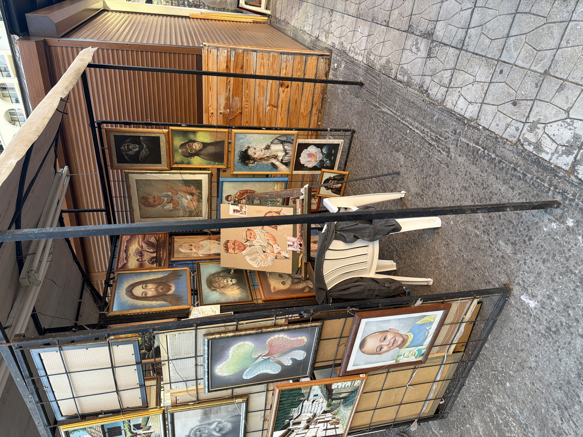 In Nesebar, Bulgaria, an outdoor art stall displays a variety of framed paintings. The stall has a metal frame supporting several pieces of art, including portraits of Jesus, children, a lion, a cat, a woman in historical attire, and monkeys. A painting of a butterfly and a still life of a rose are also visible. The stall is situated on a paved walkway with a concrete border. A white plastic chair is placed in front of the stall, with a dark jacket draped over it. The background features a building with wooden siding and a retractable awning.