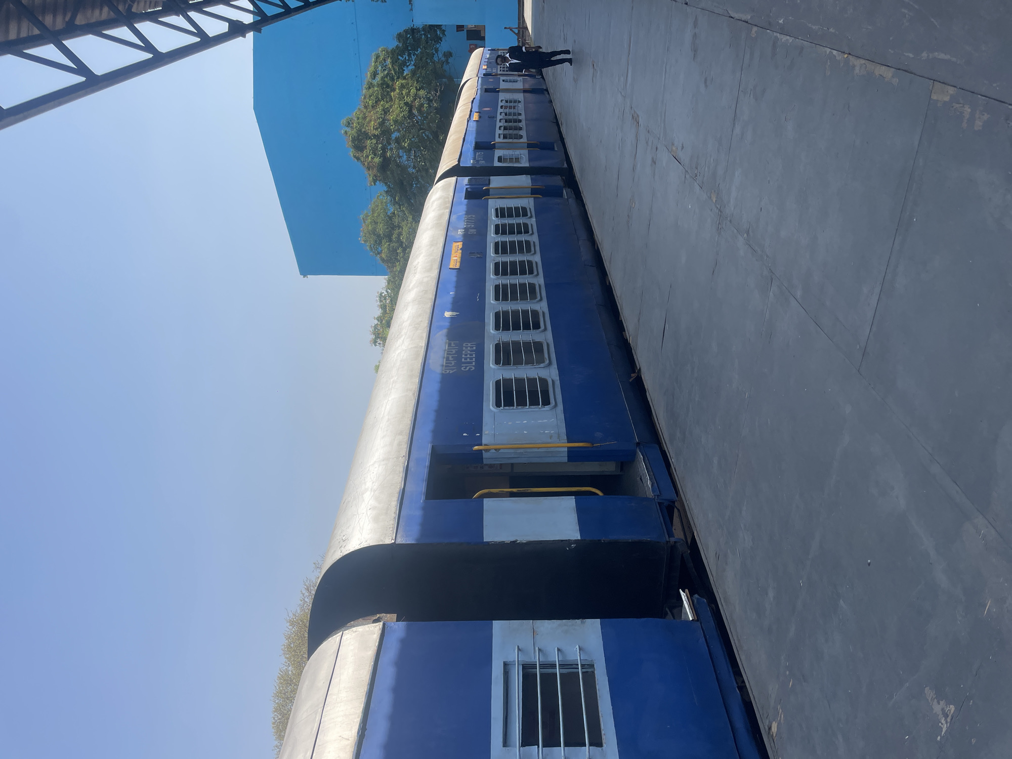 The image shows a blue and gray Indian Railways sleeper car parked on a platform in Anajpur, India. The train car has windows with bars and the word "SLEEPER" written in Hindi. A person in dark clothing is standing further down the platform near the train. A blue building and trees are visible in the background. A metal structure is overhead in the foreground. The platform is gray. The sky is blue.