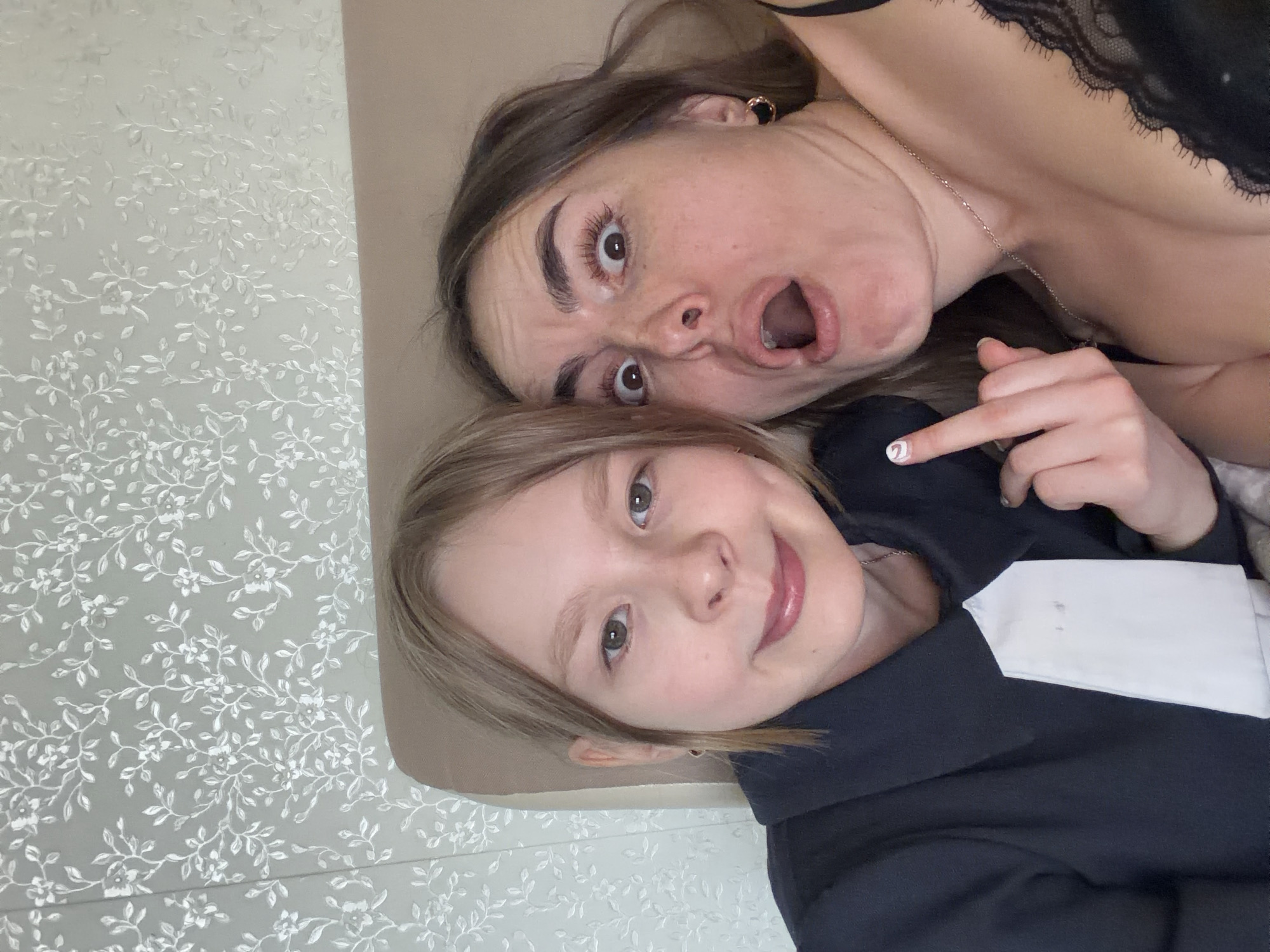 Two individuals are present in the image, a woman and a child, both lying down. The woman has brown hair, fair skin, and is wearing a black lacy top with a silver necklace. Her mouth is open in an exaggerated expression, and her eyes are wide. The child, with blonde hair, is wearing a dark jacket with a white collar and is smiling. One of the child's fingers is visible, showing a decorated nail. The background consists of a patterned wallpaper with a floral design in white and gray. There is no indication that this image was taken in Izhevsk, Russia.