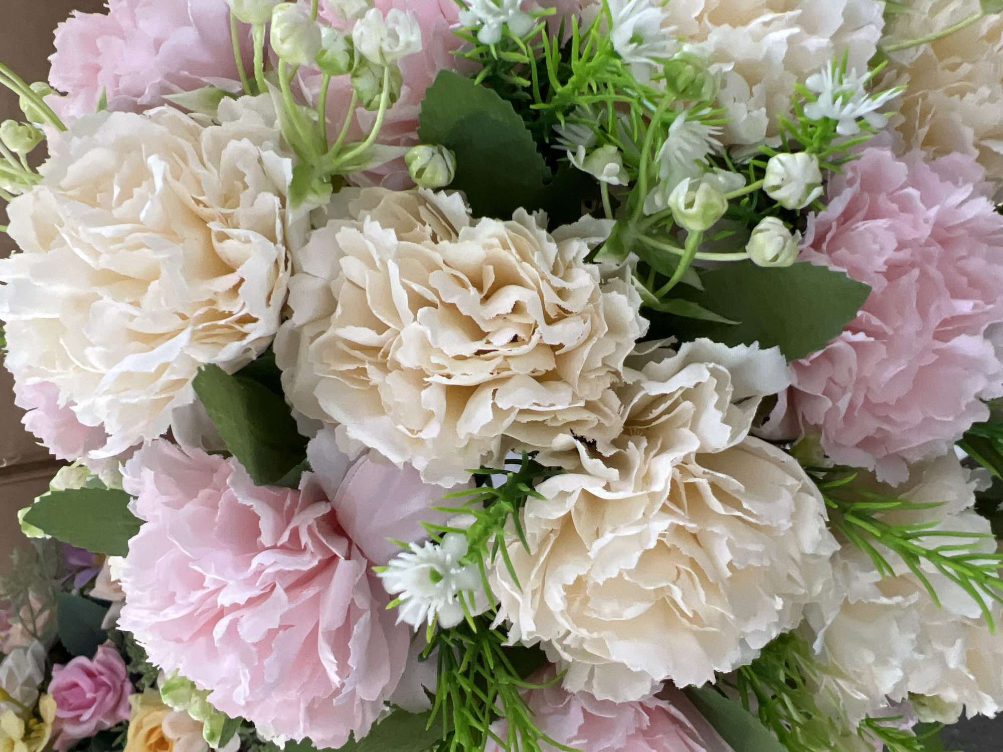 A close-up, eye-level shot showcases a vibrant bouquet of artificial flowers. The bouquet is dominated by large, fluffy blooms in shades of light pink and cream, with several smaller white flowers interspersed throughout. Bright green artificial leaves and thin sprigs add a touch of greenery. The background is blurred, providing no context for the location. The image does not depict any people or actions. There is no information available to associate this image with Eshanguzar, Uzbekistan or any other specific location in a city.