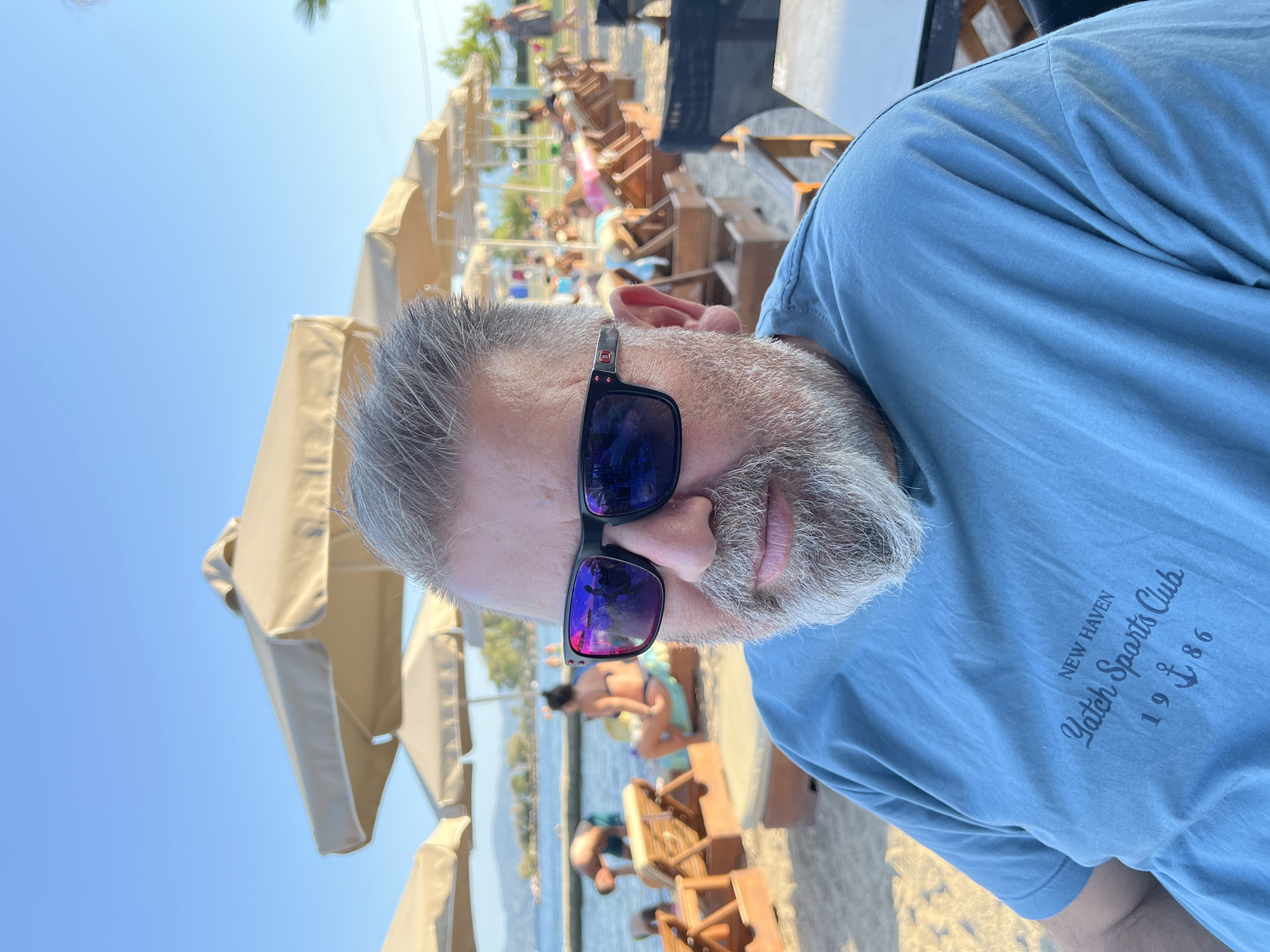 A medium shot captures a man with graying hair and a beard wearing sunglasses and a blue t-shirt. He is seated on a beach with lounge chairs and umbrellas visible in the background, along with people relaxing and the sea. The man's t-shirt features a graphic print that reads "NEW HAVEN Yatch Sports Club 1986" with an anchor symbol. The scene appears to be set during the day, with a clear blue sky. The location is a beach in the Eretria Municipal Unit, Greece.