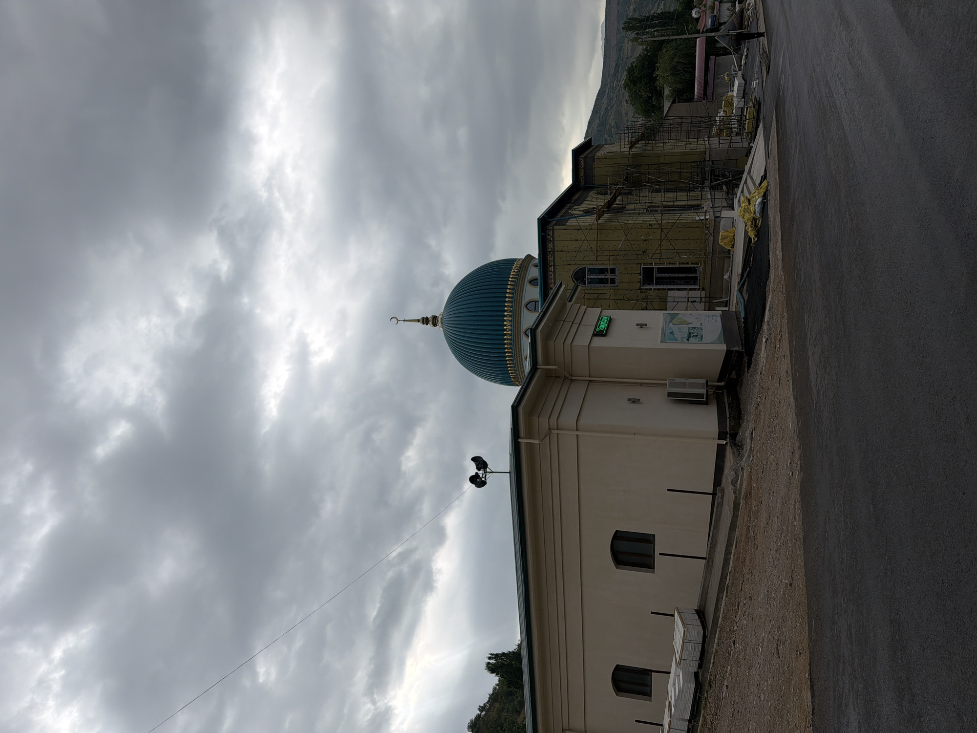 In the city of Khumsan, Uzbekistan, under a dramatic, overcast sky, a mosque is visible. The mosque features a prominent, teal-colored dome topped with a crescent moon. To the left of the dome, a cream-colored building with arched windows is situated. In front of this building, stacks of white materials are piled on the ground. Adjacent to the mosque's dome, a structure is undergoing construction, evidenced by scaffolding and insulation. A street runs across the foreground of the image, and a lone figure, carrying a large object on their back, walks away from the mosque on the right side of the frame. The surrounding environment appears hilly, with trees dotting the landscape in the background. A pair of loudspeakers are mounted on a pole atop the cream-colored building.