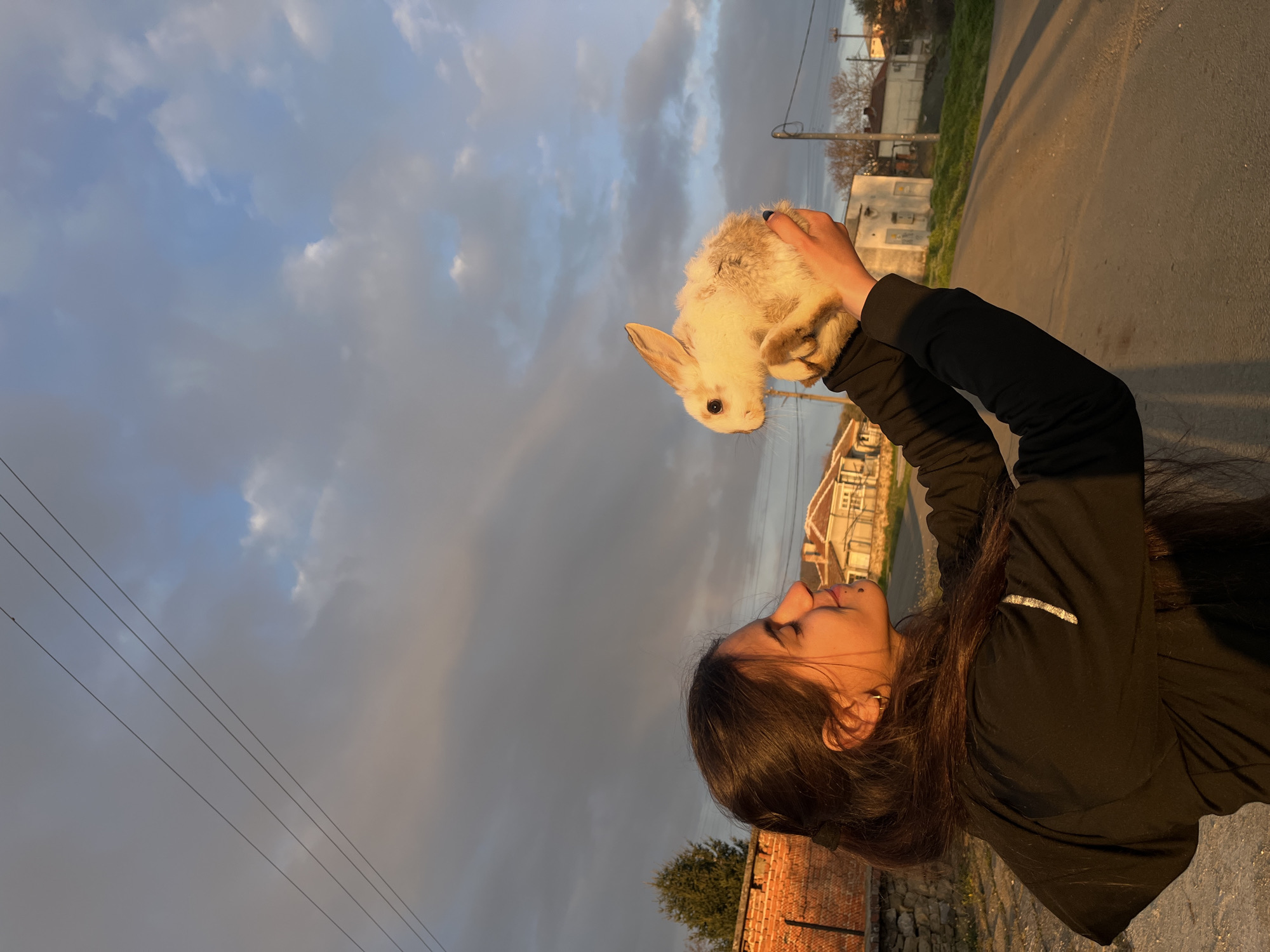 A young woman holds a small, white rabbit in her outstretched hands, gazing at it. The setting is outdoors, likely near or in the city of Lozarevo, Bulgaria, as evidenced by the typical architecture of the residential buildings and surrounding environment. The woman has long, dark hair and wears a black jacket. The rabbit appears fluffy. The background includes a road, houses, and a cloudy sky. The lighting suggests it's near sunset or sunrise. Power lines are visible in the upper left corner. A brick wall is located in the lower left corner.