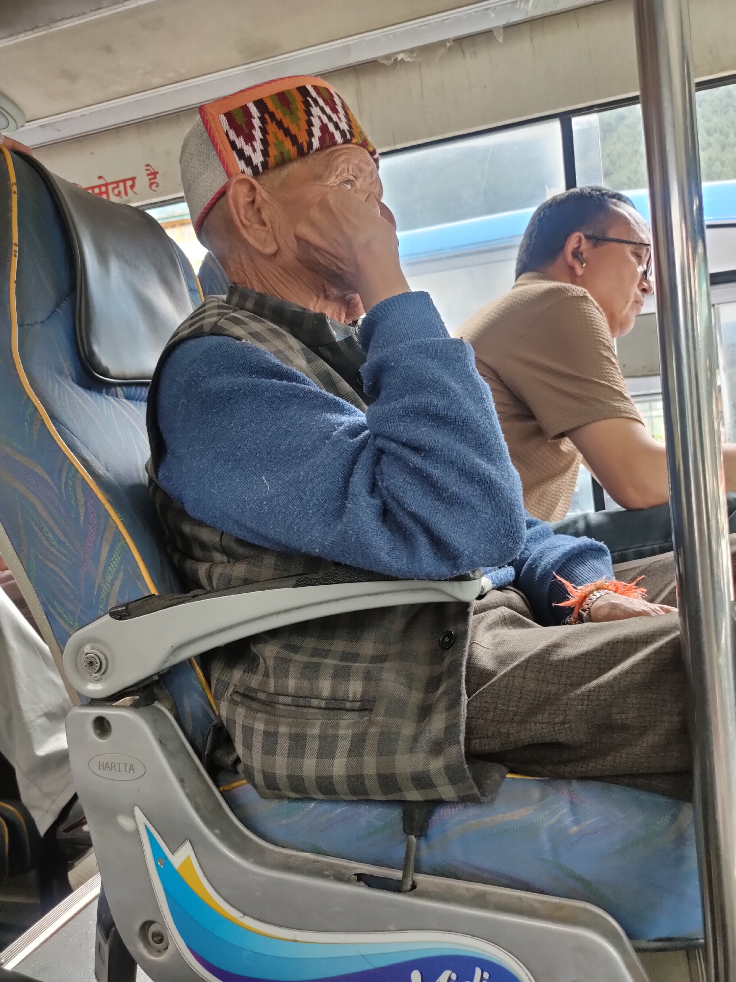 The image depicts the interior of a bus, likely in Manali, India, given the context provided. An elderly man with wrinkled skin is seated on a blue bus seat. He is wearing a traditional Himachali cap with colorful geometric patterns in red, white, green, and orange. His attire consists of a blue sweater and a grey plaid vest. He is resting his hand on his cheek, looking towards the right. In the background, another man wearing glasses and a brown shirt is also seated. The bus has large windows, revealing a glimpse of a green, tree-covered slope outside. The seats are blue with a patterned fabric. The armrest of the seat in the foreground displays the "HARITA" logo and a colorful graphic design. The overall scene suggests a common mode of public transportation in the region.