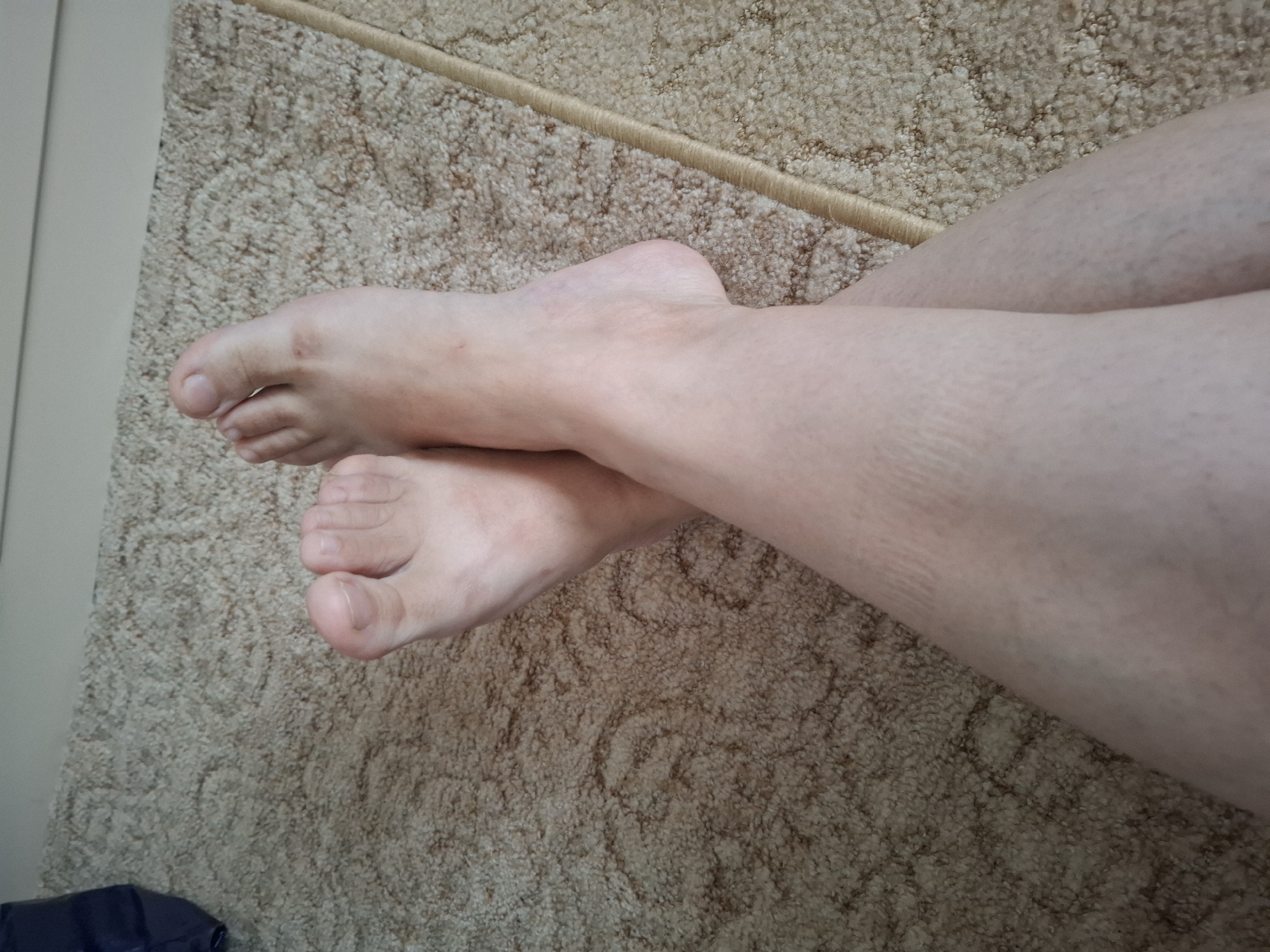 The image shows a close-up view of two bare feet, crossed at the ankles, resting on a beige, textured carpet. The feet appear to be fair-skinned and are positioned in the center of the frame. The carpet has a subtle, repeating pattern. In the upper left corner, a portion of a dark blue object, possibly a bag, is visible. The background is out of focus, but suggests a light-colored wall or furniture. The image does not provide any information about the location in the city of Chishki, Russia.