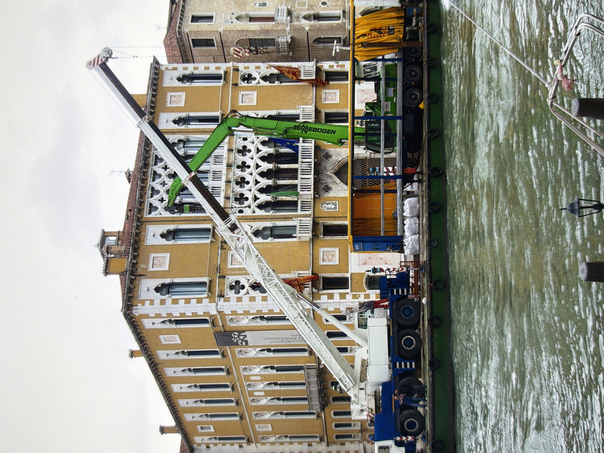The image depicts a construction scene on a canal, with a large crane positioned on a barge in front of a historic building. The building, characterized by its yellow facade, intricate white decorative elements, and arched windows, appears to be located in a European city. The crane, a substantial piece of machinery with a long white boom, is extended upwards and to the right. A smaller green crane is also visible, mounted on a truck. Several individuals are present on the barge, seemingly engaged in operational tasks. White sandbags are stacked on the barge, suggesting ongoing construction or preparation. The water in the canal is choppy and greenish, indicating a waterway in an urban setting. The sky is overcast, contributing to a muted lighting condition. The specific location in Kyiv, Ukraine, cannot be determined from the visual information provided; the architectural style is more characteristic of Italian cities like Venice.