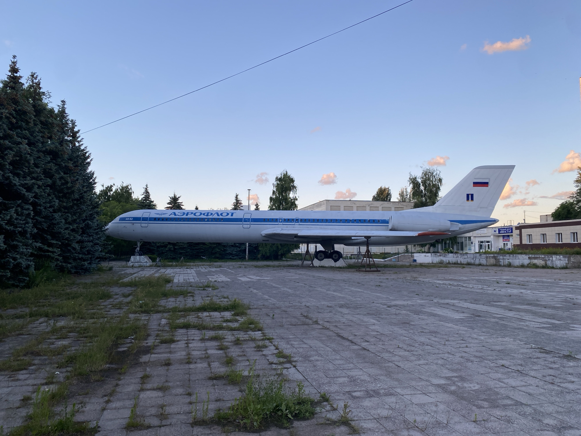 A static, full-body side view of a Tupolev Tu-154 airplane, painted white with blue accents and the word "АЭРОФЛОТ" (Aeroflot) in blue Cyrillic letters on its fuselage, is displayed on a paved outdoor area. The aircraft, bearing the Russian flag on its tail, is mounted on stands, with its landing gear visible. The scene is set in Ulyanovsk, Russia, outside the Ulyanovsk Civil Aviation Institute, where the plane serves as a monument and training aid. The surrounding environment consists of a cracked concrete or paving stone ground with patches of grass and weeds, mature evergreen trees to the left, and low-rise buildings in the background. The sky above is a clear, pale blue with a few wispy clouds. The lighting suggests it is either early morning or late afternoon. No people are visible in the frame.