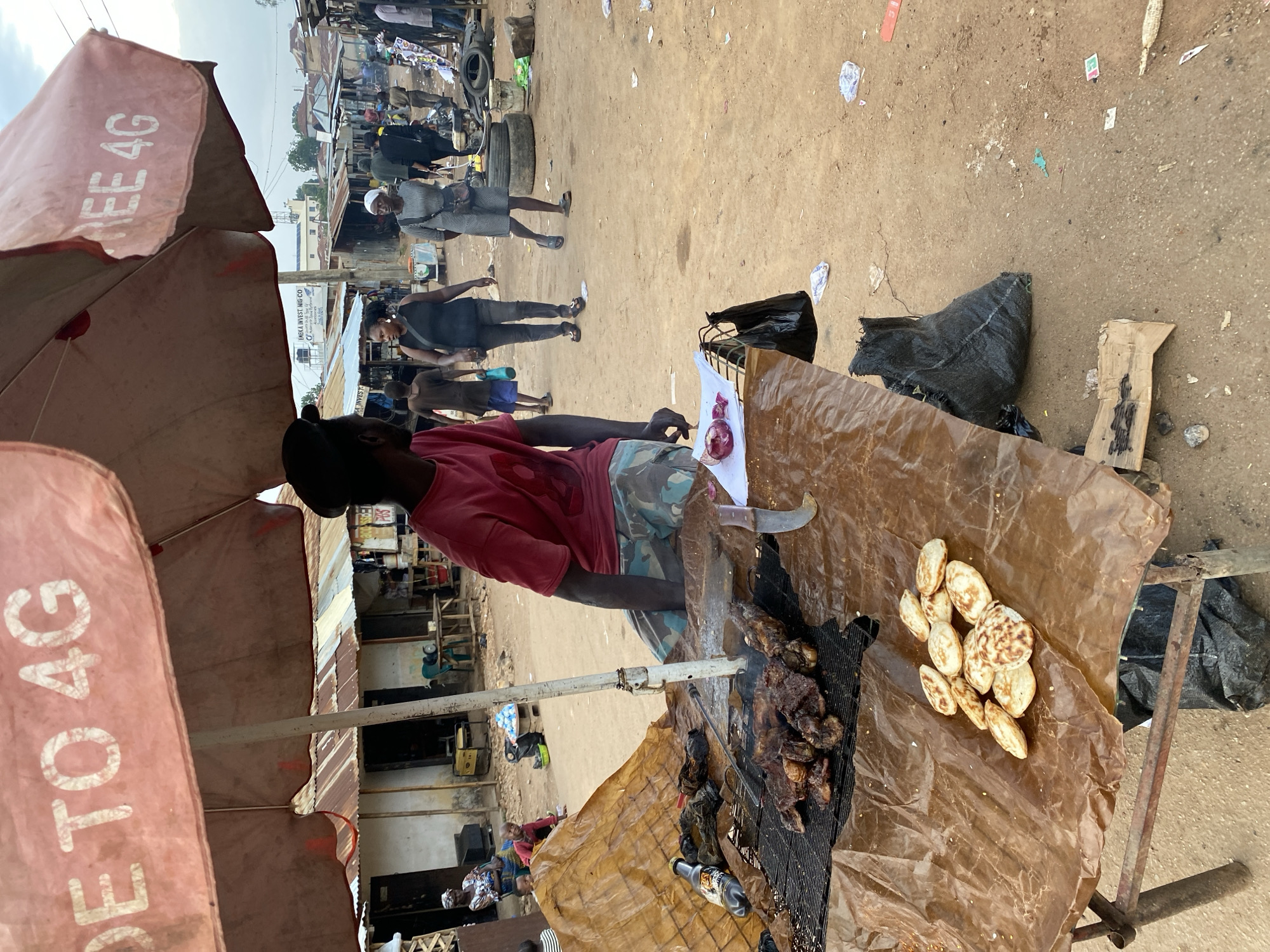 In Bukuru, Nigeria, a man in a red t-shirt and camouflage shorts sits under a large, faded pink umbrella advertising "4G" internet. He is positioned behind a makeshift food stall, where grilled meat is cooking on a wire rack over coals. A brown tarp covers the table, and several small, round fried dough items are arranged on it. A bottle of drink and a knife with a curved blade are also on the table. In the background, people walk through a dusty market, with various stalls and corrugated metal structures visible. The sky is overcast, suggesting a potentially humid or rainy day. The scene captures a moment of daily commerce in a bustling Nigerian marketplace.