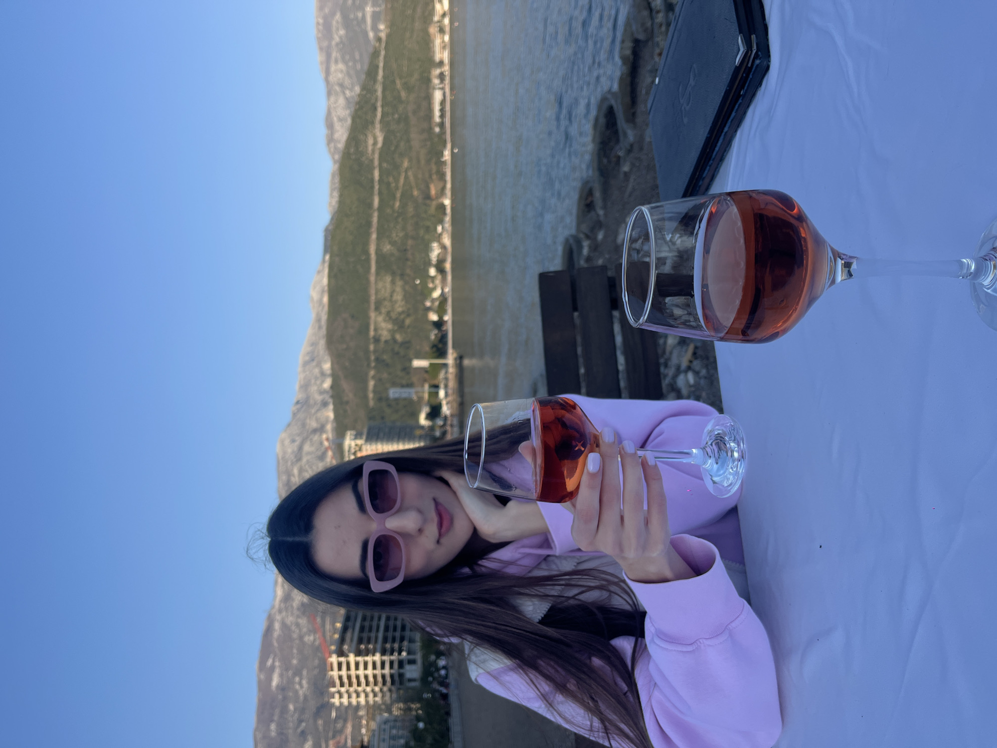 A young woman wearing pink sunglasses and a pink sweatshirt sits at a table with a white tablecloth outdoors, holding a glass of rosé wine. A second glass of rosé wine sits on the table to her right, next to a dark-colored menu. In the background is the city of Budva, Montenegro. The location appears to be a restaurant or bar overlooking the Adriatic Sea, with mountains visible in the distance. The sky is clear blue. The image is rotated 90 degrees counterclockwise.