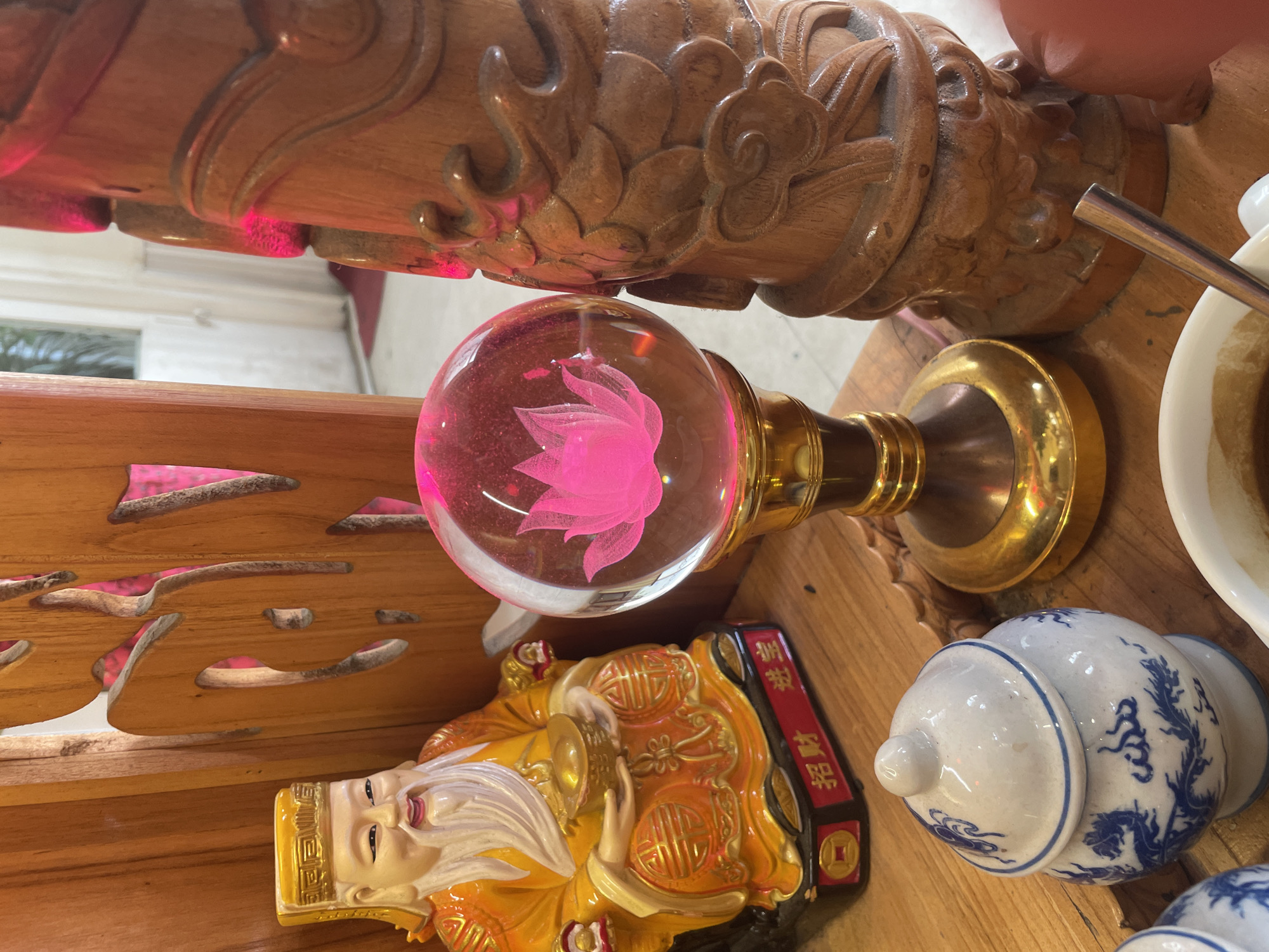The image displays a collection of religious and decorative items on a wooden surface, likely in a temple or shrine in Nha Trang Ward, Vietnam. On the left, a figurine of a seated elder, possibly a deity or revered figure, is dressed in an orange robe with gold trim and holds a gold ingot. The base of the figurine has Chinese characters. Beside it, a clear glass sphere with a pink lotus flower motif inside sits on a golden stand. To the right, a carved wooden pillar with intricate dragon or cloud motifs is visible. In the foreground, a small ceramic jar with blue dragon designs and a lid is placed on the wooden surface, along with parts of what appears to be a white bowl containing a brown liquid and a metal utensil. The background is slightly blurred, showing wooden structures and hints of an interior space.