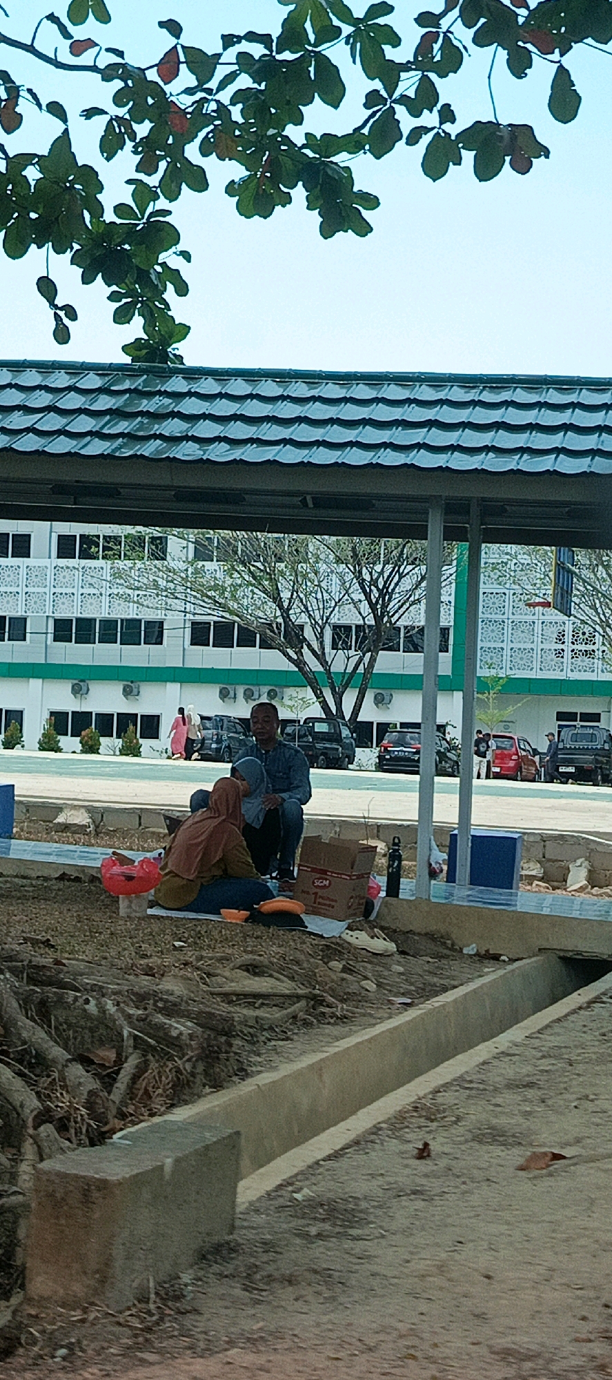 In Desa Namang, Indonesia, a man and a woman are seated on the ground, facing each other, with food items spread out on a mat between them. The man, wearing a blue collared shirt and dark pants, is sitting upright. The woman, clad in a headscarf and a brown hijab with a blue and green garment, is leaning towards the man. Several objects are visible: a red plastic container, a cardboard box, a black water bottle, and a pair of white shoes. In the background, a modern building with white walls and a green tiled roof stands behind a line of parked vehicles including black pick-up trucks, a red car, and a black SUV. The environment appears to be an outdoor seating area with exposed tree roots and a concrete drainage channel in the foreground. The sky is a pale blue.