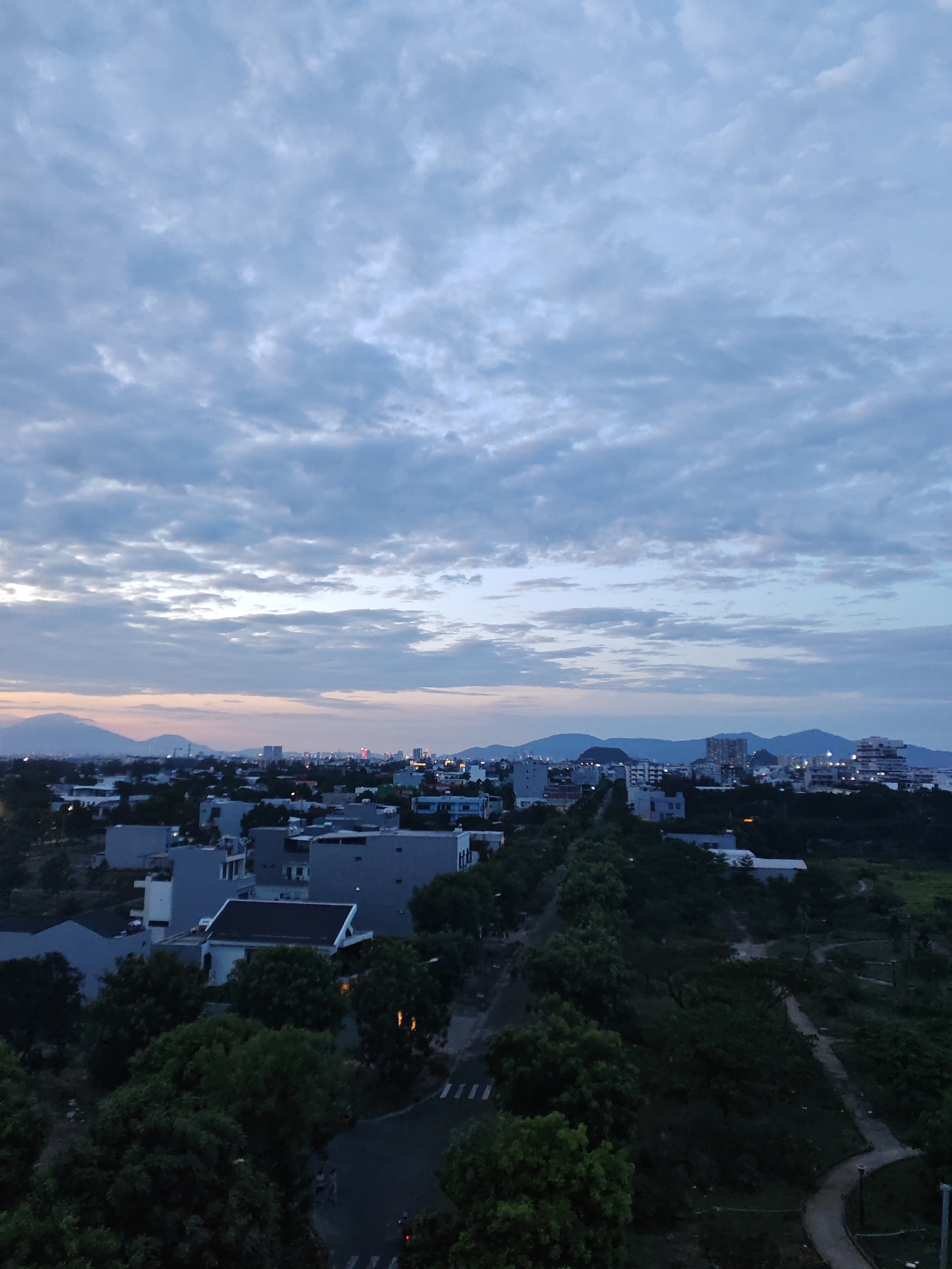 The image depicts a panoramic view of a city at dusk, likely in Đà Nẵng, Vietnam, given the presence of coastal mountains characteristic of the region and the architectural style of the buildings. The sky is filled with a dramatic expanse of clouds, tinged with the soft colors of sunset. Below the sky, a cityscape unfolds, characterized by a mix of low-rise residential buildings and taller structures in the distance. Dense greenery, primarily trees, covers a significant portion of the foreground and middle ground, suggesting a city with ample parks or natural landscapes interspersed with urban development. A road winds through the scene, with some streetlights visible, indicating the presence of infrastructure. In the distance, mountains form a hazy backdrop against the horizon. The overall atmosphere is serene and tranquil, capturing a moment of transition between day and night.