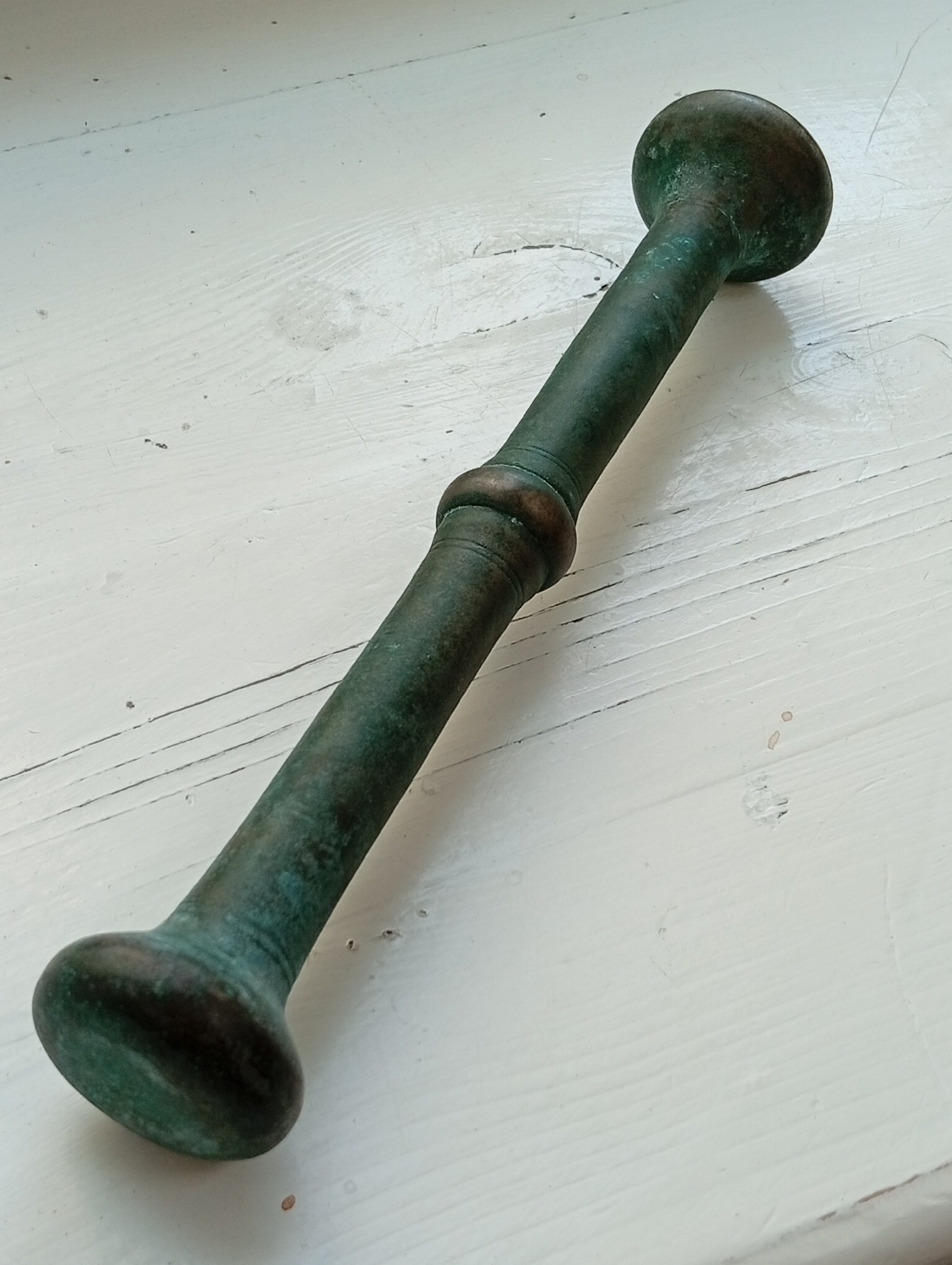 A tarnished bronze mortar and pestle rests on a white wooden surface. The mortar has a bulbous end and a long, slender shaft that tapers to another bulbous end. A decorative band circles the shaft in the middle. The surface beneath is made of planks with visible wood grain. The scene appears to be indoors, possibly on a windowsill or table, given the lighting and shadows. No people or specific locations within Крыўлянскі сельскі Савет are identifiable from the image alone.