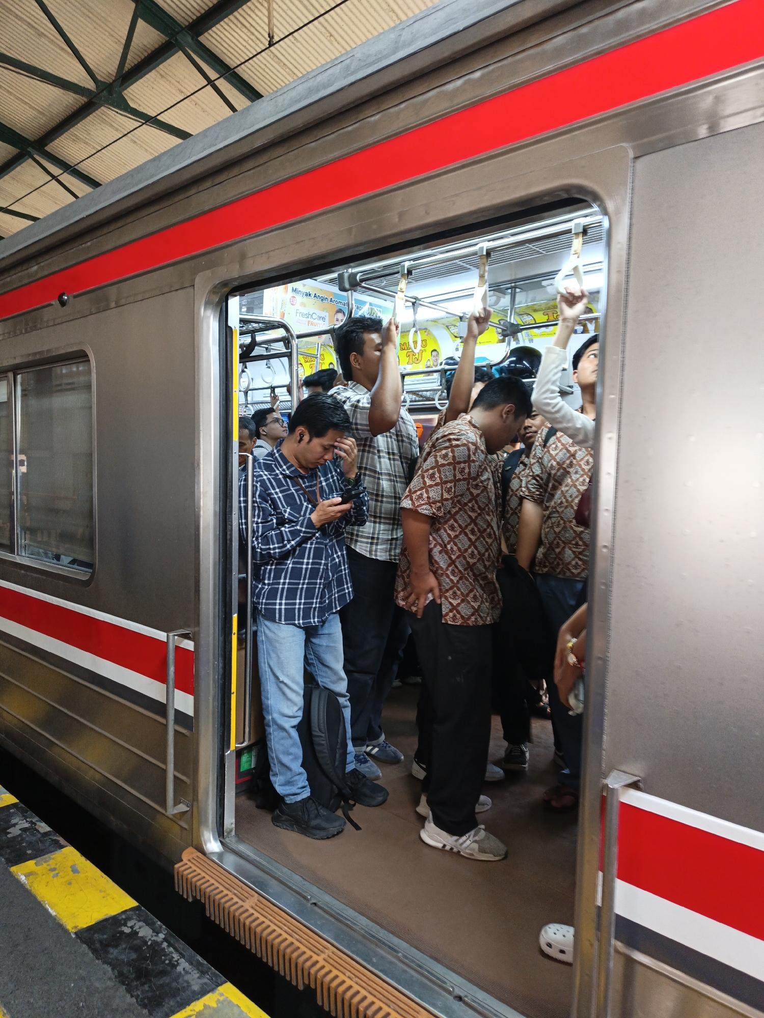 This image depicts the interior of a crowded commuter train car in Klaten, Indonesia. Several individuals are standing, some holding onto overhead handrails. A man in a blue checkered shirt and jeans is looking at his phone, while another man in a patterned brown shirt is also visible. The train car appears to be a typical urban transit vehicle, with metal walls, handrails, and advertisements visible on the interior. The lighting suggests it is daytime. The platform outside the train is partially visible, marked with yellow and black lines. The scene captures a moment of daily commute on Indonesian public transportation.