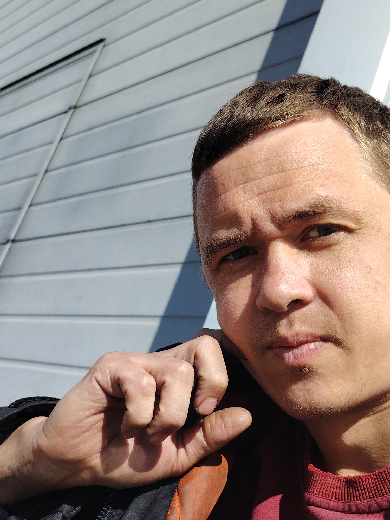 A medium close-up photograph shows a fair-skinned man with short brown hair, gazing directly at the camera with a neutral expression. His right hand is raised and positioned near his cheek. He appears to be wearing a maroon shirt with a ribbed collar, partially covered by a black jacket with an orange lining. The background consists of a light gray building with horizontal siding. The location is Noviy, Russia.