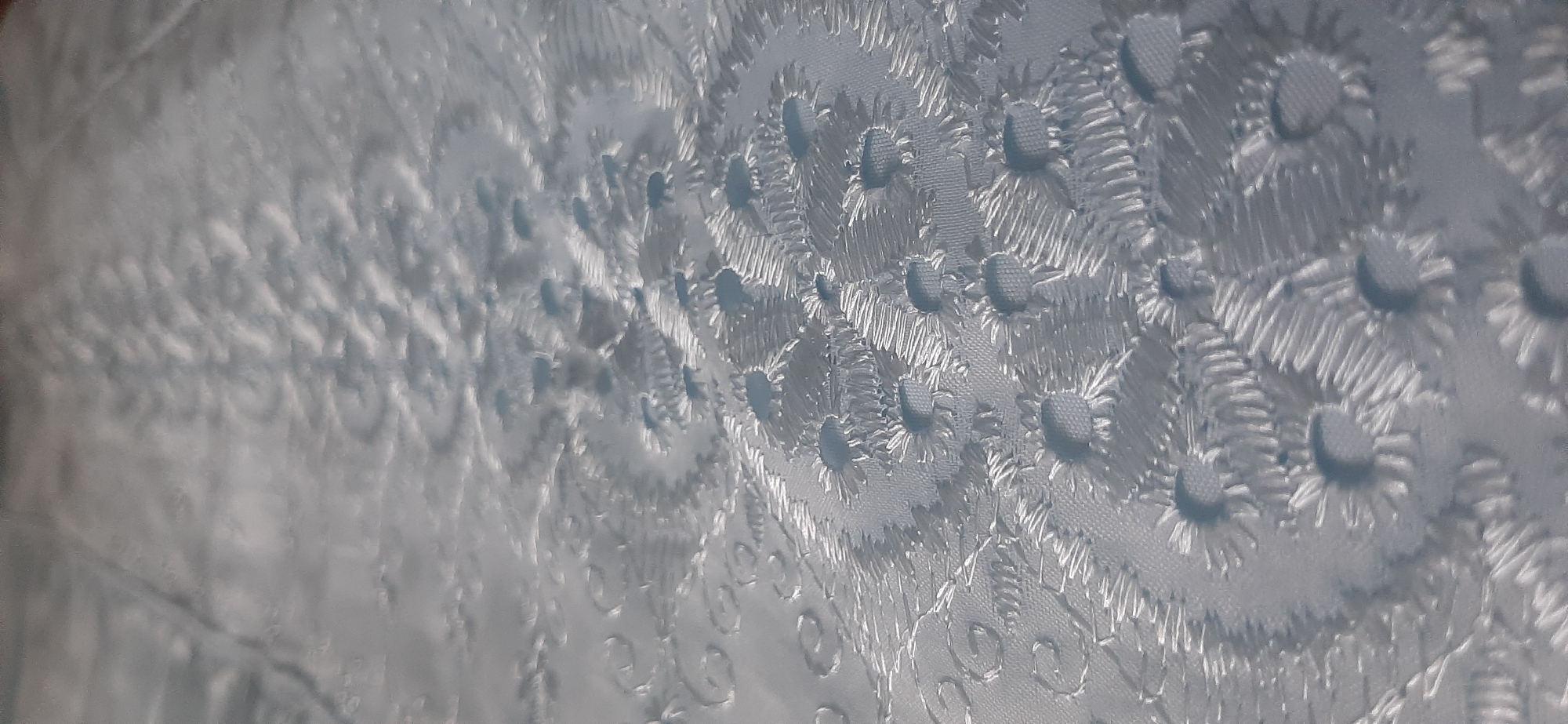 The image displays a close-up, high-angle shot of a light blue fabric with an intricate, raised floral pattern. The fabric appears to be a textured material, possibly a brocade or jacquard weave, with the floral motifs appearing to be embroidered or woven in a slightly darker shade or with a different sheen, creating a subtle contrast. The lighting emphasizes the three-dimensional quality of the pattern, with shadows highlighting the raised elements. The background is a soft blur, indicating a shallow depth of field. There are no people, animals, or discernable objects other than the fabric itself. The image does not contain any information to determine its location in Yessentukskaya, Russia, or any other city.