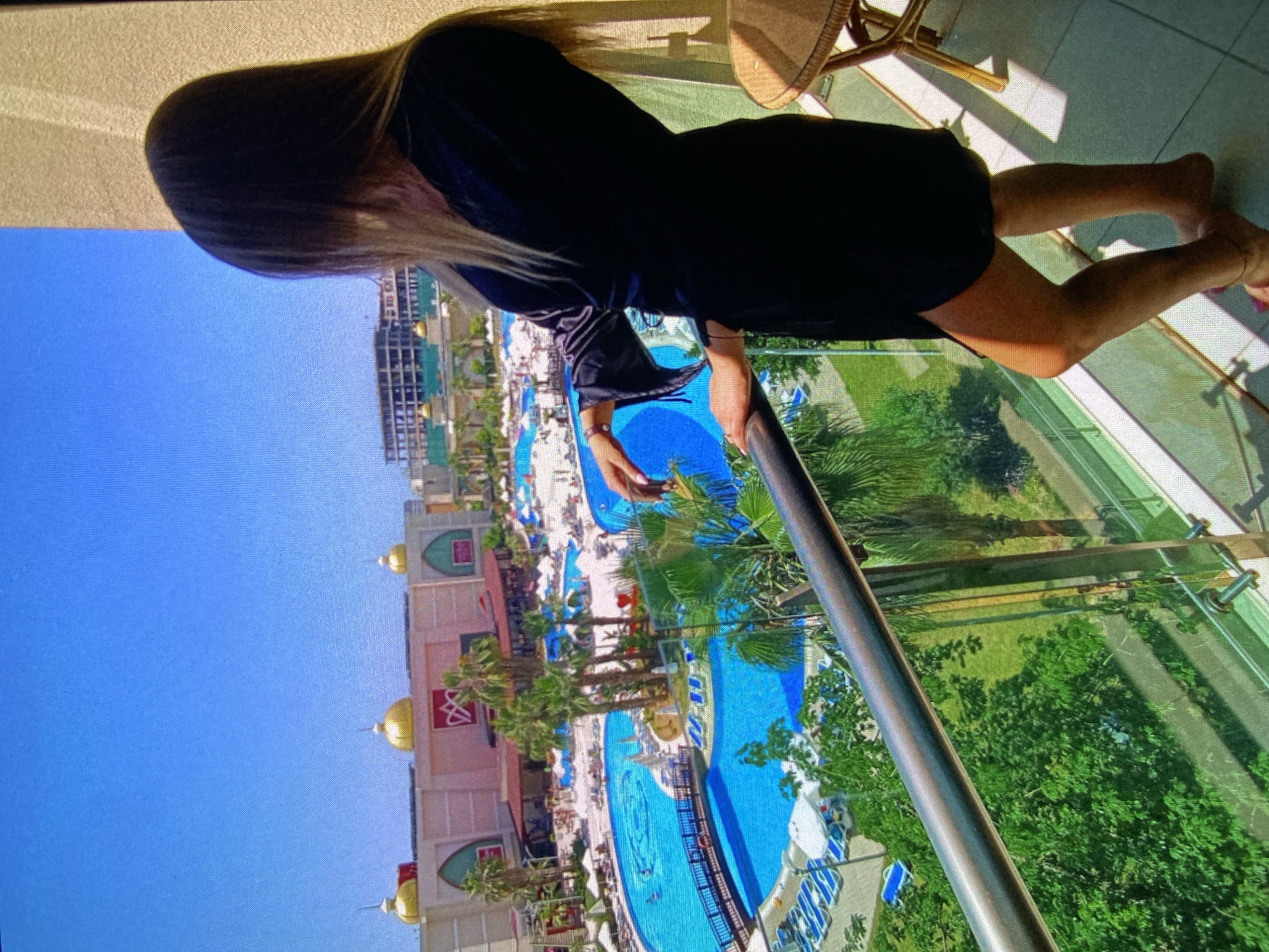 A high-angle shot captures a person standing on a balcony, observing a large resort complex below on a clear, sunny day. The individual, with long, light-colored hair and dressed in dark attire, faces away from the viewer, hands resting on the metal railing.
Below the balcony, the expansive resort features multiple brightly-lit blue swimming pools of varying shapes, surrounded by numerous sun loungers and scattered palm trees. Small figures are discernible in and around the pools, indicating activity. The resort buildings are light-colored, adorned with distinctive golden dome-shaped roofs and other architectural details. On one building facade, a red logo resembling intertwined 'M's or a stylized 'W' is visible against a white background. Beyond the resort, lush green foliage and other distant buildings complete the panorama.
The time of day appears to be mid-day or early afternoon, marked by the brilliant blue sky and strong sunlight casting distinct shadows. The scene evokes a vacation or leisure setting in Bankya, Bulgaria.