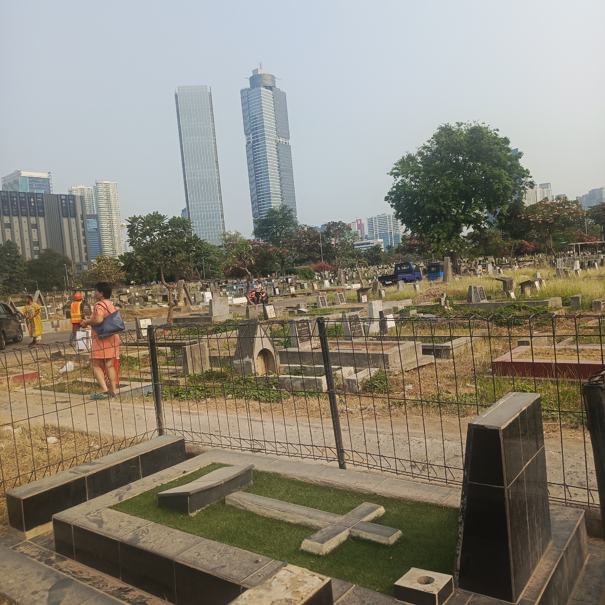 In the foreground, a grave marker is visible with a dark stone border surrounding a patch of artificial turf. A cross-shaped stone marker lies on the turf. Behind this grave, a black metal fence separates it from a larger cemetery area. The cemetery contains numerous gravestones and mausoleums, some appearing old and weathered, others more modern. In the middle ground, three people are visible. One, wearing an orange vest and hard hat, appears to be a worker, possibly tending to the cemetery. Another person in a yellow garment stands near a parked car. A third person, a woman in a salmon-colored dress, walks away from the camera carrying a dark blue tote bag. Further back, in the background, a dense urban landscape is visible, characterized by several tall, modern skyscrapers. The sky is hazy, suggesting a warm, possibly humid day in the city. This scene likely depicts a cemetery juxtaposed with a modern cityscape in South Jakarta, Indonesia, a common sight in densely populated urban areas where historical sites coexist with contemporary development.