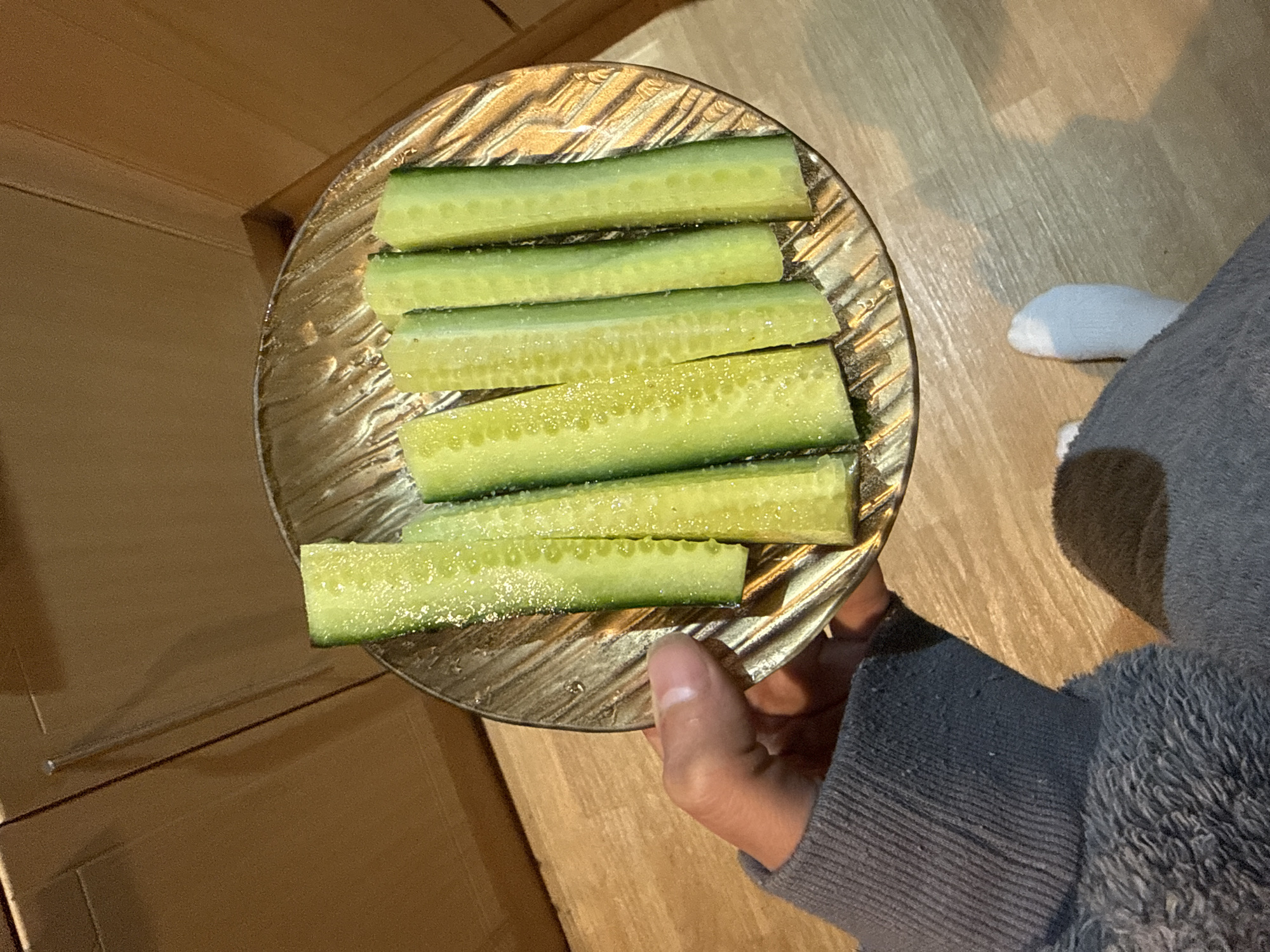 A person holds a clear, textured glass plate with six sliced cucumbers in their left hand. The cucumber slices are arranged in parallel rows, glistening with moisture, possibly water or oil. The person is wearing a gray, long-sleeved garment. A white sock peeks out from the garment, suggesting the lower leg of the person. The background shows a wooden surface, with cabinets or drawers on the left and a wood floor on the right. No information suggests this image was taken in Great Yarmouth, United Kingdom.