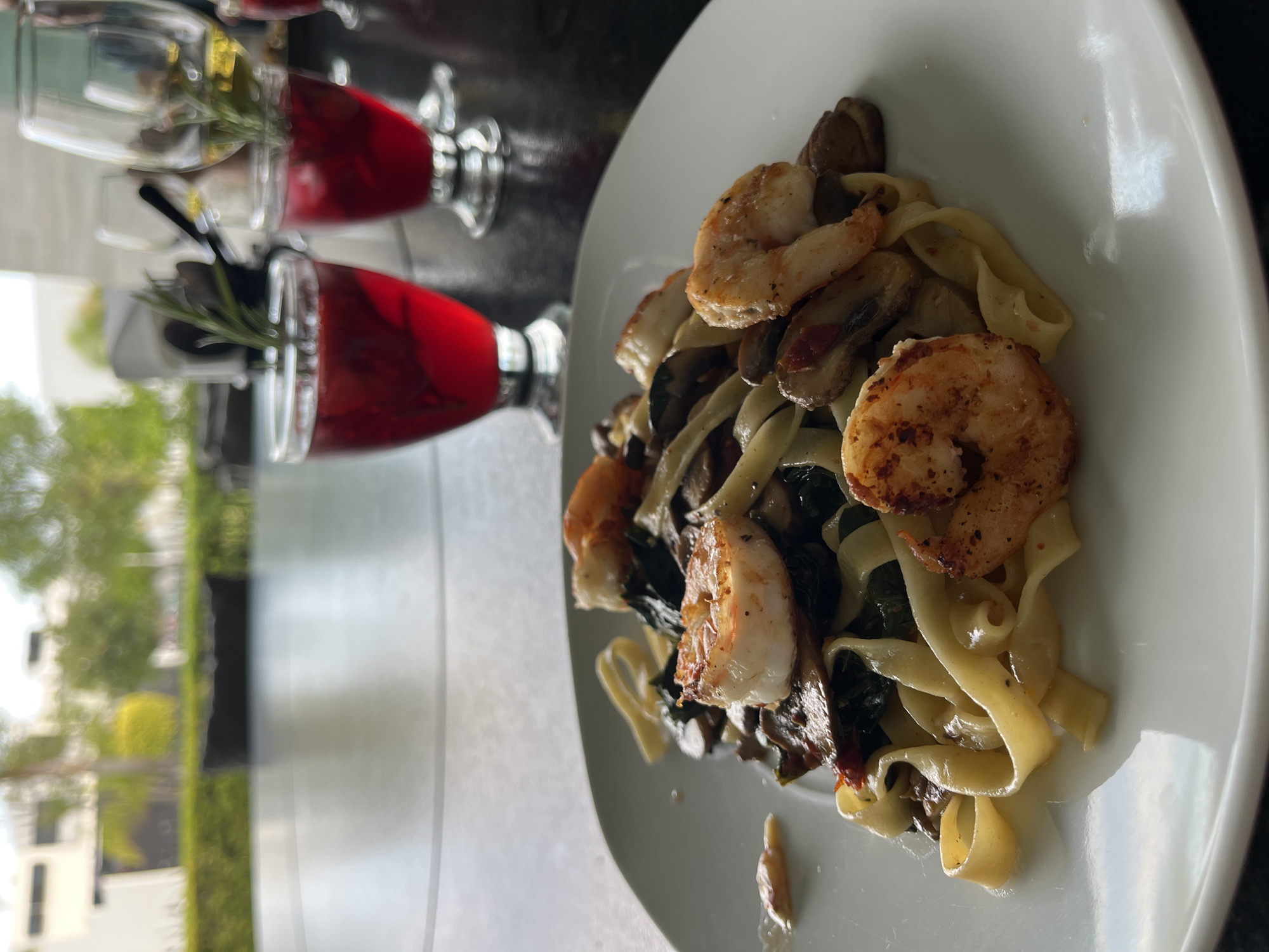A close-up shot shows a plate of pasta with shrimp and mushrooms next to two glasses of red liquid with rosemary sprigs in San Bernardino Tlaxcalancingo, Mexico. The pasta appears to be fettuccine or a similar wide noodle. The shrimp are arranged on top of the pasta, exhibiting a cooked appearance with some browning. Sliced mushrooms and dark green leafy vegetables are also mixed in with the pasta. The plate is white. The glasses contain a dark red liquid and are garnished with a sprig of rosemary. The background includes a blurry view of trees and buildings, suggesting an outdoor or window-side setting. The lighting is bright.