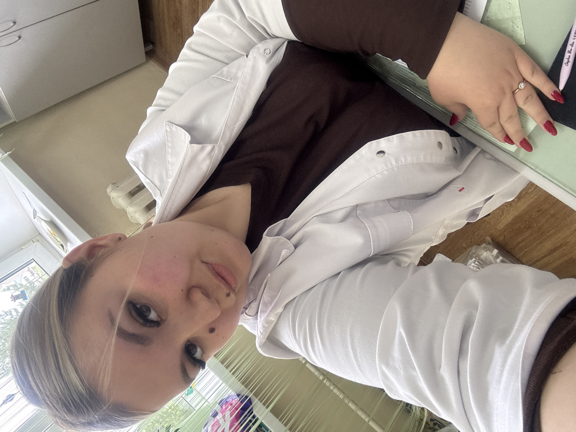 A young woman with fair skin, blonde-streaked hair, and dark eyes is posing for a selfie indoors. She is wearing a white lab coat over a brown top. She has a ring on her left ring finger and red nail polish. The background includes a window with visible greenery, a white cabinet, a radiator, and papers on a desk. It is not possible to confirm the location as the city of Фоки, Russia from the image alone.