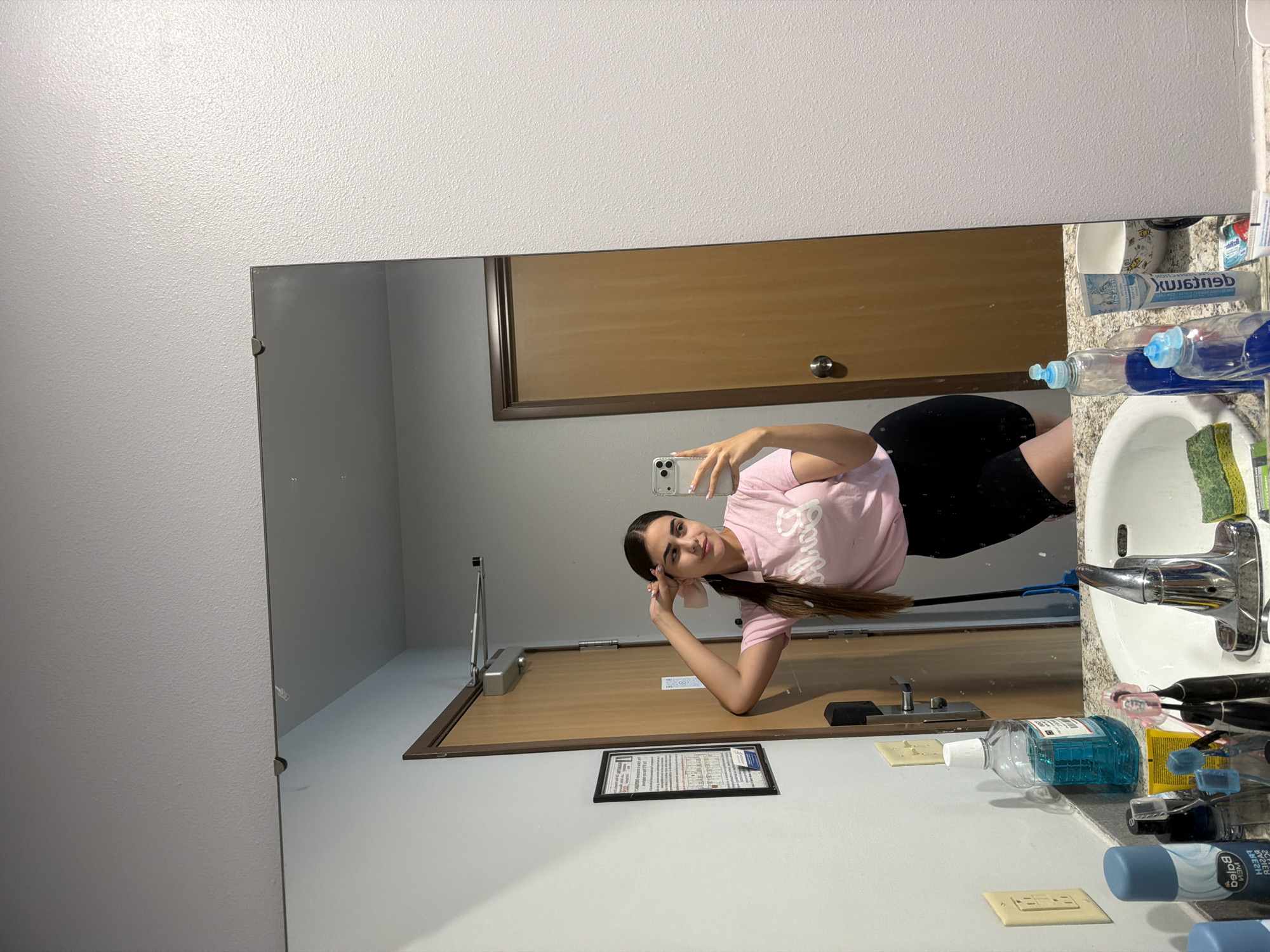 A young woman with dark hair pulled into a ponytail stands in a bathroom and takes a selfie in a mirror. She is wearing a pink t-shirt with "Barbie" written in white lettering and black shorts. She is leaning against a wooden door and has her left hand behind her head, holding her hair. A mop with a blue handle is leaning against the door. In the foreground, a white sink is visible with various toiletries on the counter, including bottles of mouthwash, a tube of toothpaste, and toothbrushes. The bathroom has light gray walls and a textured white ceiling. A framed notice is attached to the wall next to the door. The overall scene appears to be a residential bathroom, possibly in Lake Delton, Wisconsin, United States of America.