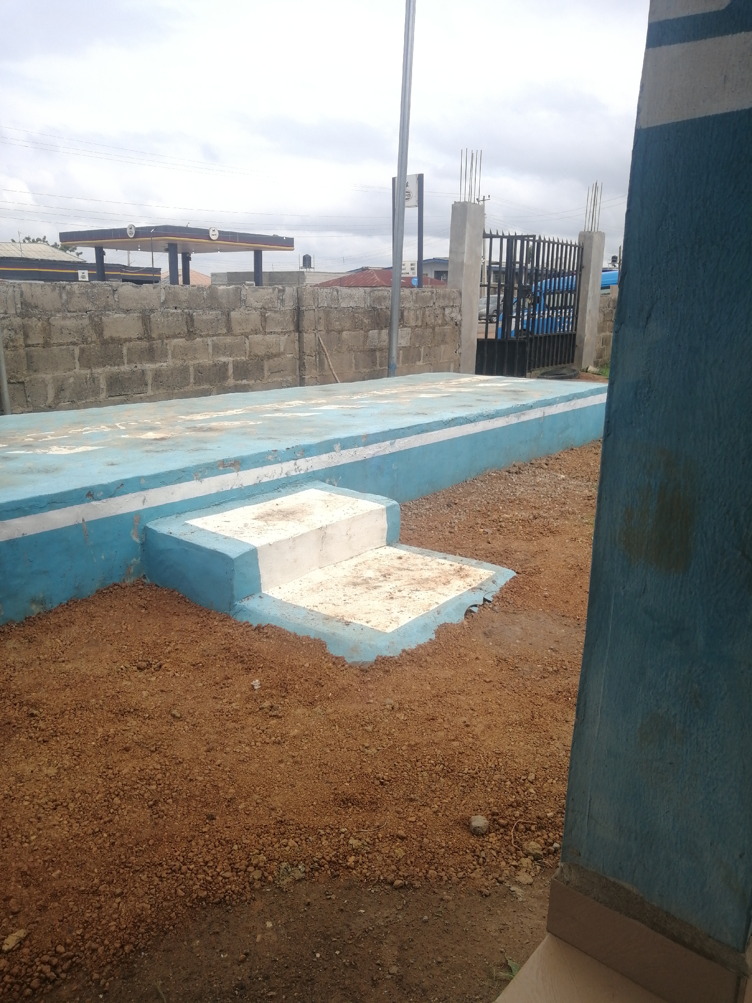 The image shows a concrete platform with blue and white painted borders and steps, situated in an outdoor environment. The platform is surrounded by reddish-brown soil. In the background, there is a brick wall and a black metal gate with vertical bars. Beyond the gate, a gas station canopy with signage is visible. The sky is overcast. The scene is located in Ajasse Ipo, Nigeria.