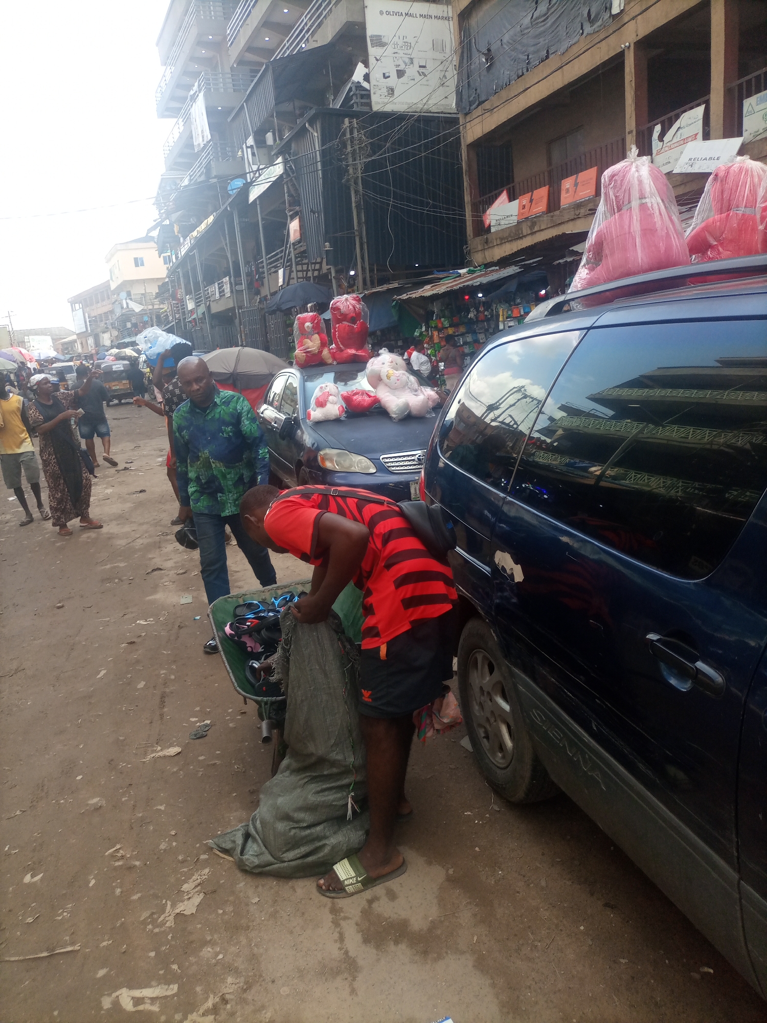 This media file captures a bustling street scene in Onitsha, Nigeria, likely during daylight hours given the bright sky and shadows. The setting appears to be a market or commercial area with multi-story buildings lining a dusty, unpaved street.
The foreground is dominated by a dark blue van, possibly a minivan, with the name "SIENNA" visible on its side. Parked alongside it, or perhaps a temporary stall, is a pushcart containing items that look like footwear and possibly some bundled goods. A man in a red and black striped shirt is actively engaged with the cart, bending over and appearing to sort or pack items. He is wearing black shorts and green sliders.
Beside the van and cart, another man in a green and blue patterned shirt stands looking towards the foreground. He is wearing blue jeans and black shoes. To the left of these men, a woman in a brown and black patterned dress and headscarf stands talking to someone out of frame. Further left, a person in a yellow t-shirt and grey shorts is visible.
In the background, the market stretches out with more people, vehicles including what appears to be a tricycle taxi, and various stalls or shops. Several umbrellas are open, suggesting either shade from the sun or protection from potential rain. Red teddy bears, some wrapped in plastic, are visible on the roof of the van and also on top of a blue car in the mid-ground, indicating merchandise being sold or transported. The overall impression is one of everyday commercial activity in a busy urban environment. Visible text includes "SIENNA" on the van and "OLIVIA MALL MAIN MARKET" on a sign in the upper part of the image.
