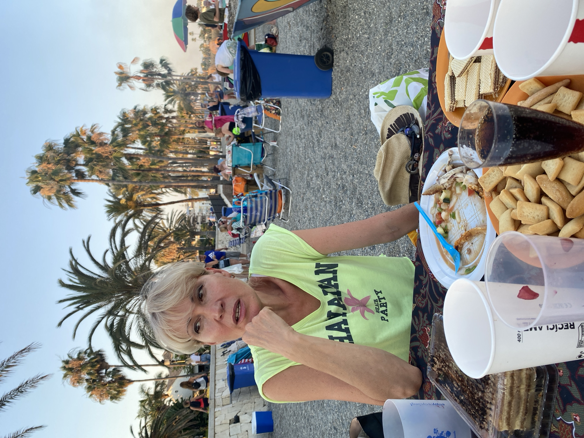 A woman with blonde hair and fair skin, wearing a yellow t-shirt with "HAWAIIAN BEACH PARTY" printed on it, is seated at a table outdoors, with a body of water and palm trees in the background. She is resting her chin on her hand, looking to her left with an engaged expression. In front of her on the table, there are various food items, including a cake, a plate of what appears to be leftovers from a meal with chicken bones, a plate of wafers, and a glass of dark liquid, likely soda. There are also several paper cups and a straw. The table is covered with a patterned cloth. Behind the table, people are visible sitting on chairs and walking, with beach umbrellas and palm trees suggesting a beachside location. The overall scene depicts an outdoor gathering or party, potentially in Marbella, Spain, given the palm trees and beach environment.