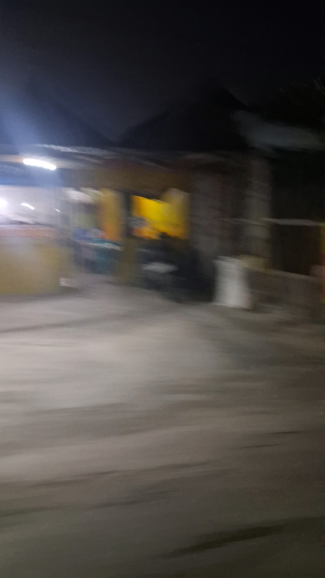 The image depicts an outdoor scene at night, likely in a semi-urban or rural area with some commercial activity. The overall image is blurry due to motion blur, suggesting it was taken from a moving vehicle.
In the background, there is a structure that appears to be a building or a stall with a roof. Bright yellow and orange elements are visible on this structure, possibly signage or lights. To the left of this structure, a bright white light source is present, possibly a streetlamp or the headlights of another vehicle.
In the mid-ground, several objects are present, including what appears to be a dark-colored vehicle or large object, and a white cylindrical object that might be a trash can or a container. The ground in the mid-ground is rough and uneven.
The foreground consists of a paved surface, likely a road or a parking area, which is also affected by motion blur. The lighting conditions are dim, with ambient light coming from artificial sources.
The location is specified as Jogotirto, Indonesia. The scene appears to be a street or an area with some businesses operating at night. There are no discernible individuals or specific actions clearly visible due to the blur. The environment suggests a typical Indonesian setting with local businesses or street vendors.