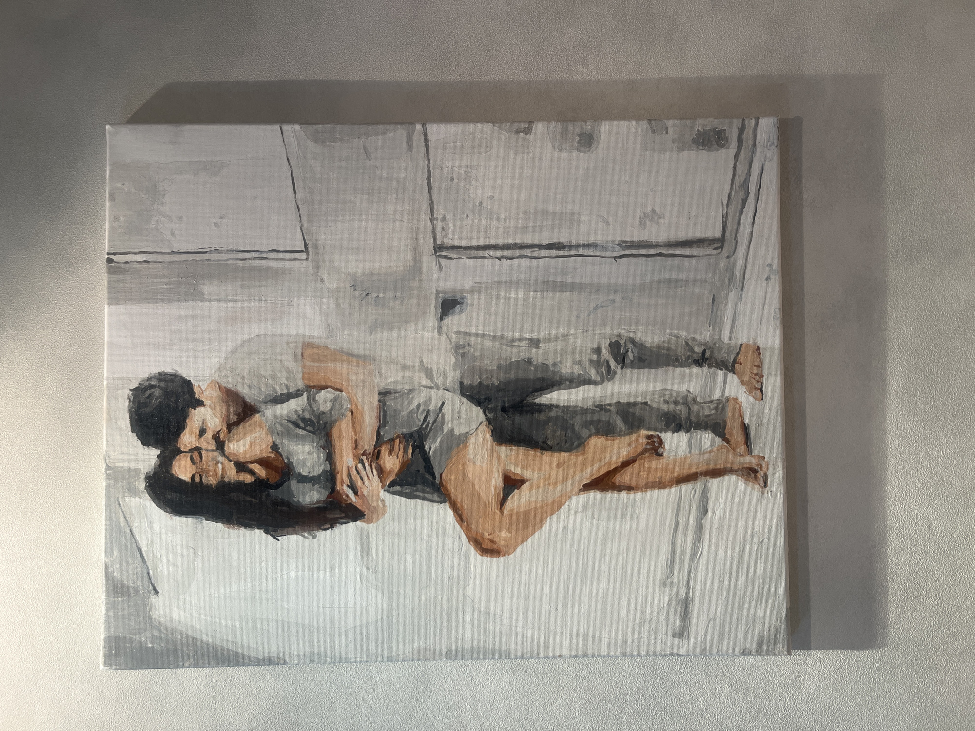 A canvas painting depicts a man and woman embracing. The woman, with long dark hair, leans back with her eyes closed, her legs crossed and one foot resting on the man's shin. She is wearing a loose gray top and possibly shorts. The man, with dark curly hair and a mustache, holds her close, his face nuzzled into her neck. He is wearing a light-colored t-shirt and gray pants. Their bodies are painted in earthy tones with hints of pink, while their clothing is rendered in shades of gray. The background is abstract, with vertical and horizontal lines suggesting an interior space, painted in muted grays and whites, with hints of light and shadow. The painting is framed and hangs on a textured, light-colored wall. This painting is located in Rybnitsa, Moldova.