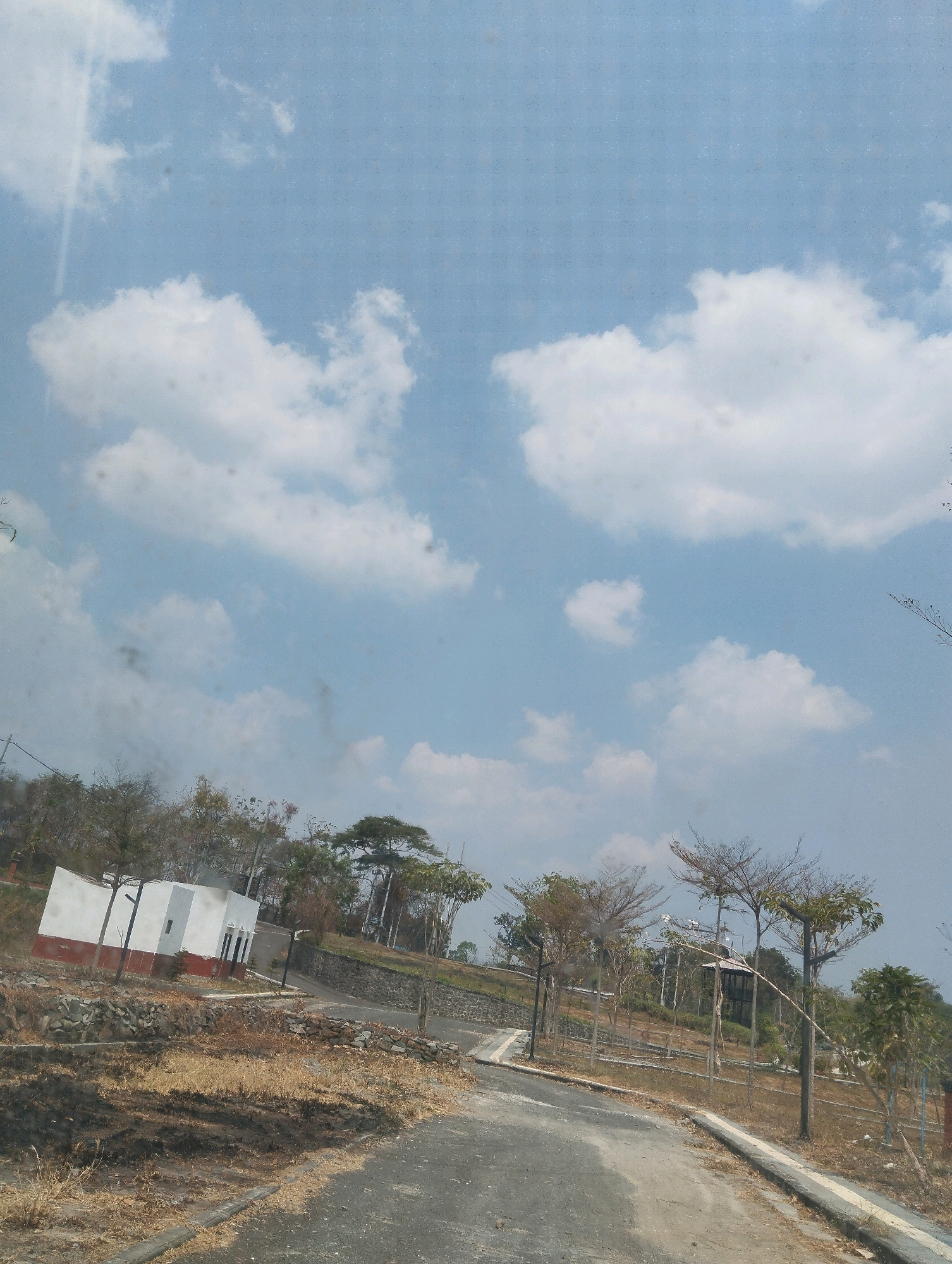 An outdoor, eye-level, vertical medium shot captures a slightly hazy, bright, daytime scene. The upper two-thirds of the frame are dominated by a pale blue sky with scattered, fluffy white clouds. A linear streak of light, possibly a reflection or lens flare, is visible on the left side of the sky.
The lower third of the image shows a dry, somewhat arid landscape. A paved, grey asphalt road curves from the bottom center towards the upper left. The road is bordered by a concrete curb and dry, yellowed grass and dirt. To the left of the road, a white building with a red-brown foundation is situated on a slope. The building has two dark-colored doors and a small rectangular window. A retaining wall made of grey stones separates the building and the road from a patch of burnt, dark earth in the foreground.
Beyond the building and road, the landscape is characterized by sparse, dry vegetation and trees. Some trees are leafless, with bare branches, while others have sparse green foliage. The terrain appears to be hilly, with more trees and a stone wall visible further up the slope. Streetlights with green lamps are positioned along the road. The overall impression is one of a developing or recently cleared area with a focus on landscaping and infrastructure. The scene is devoid of people or animals.