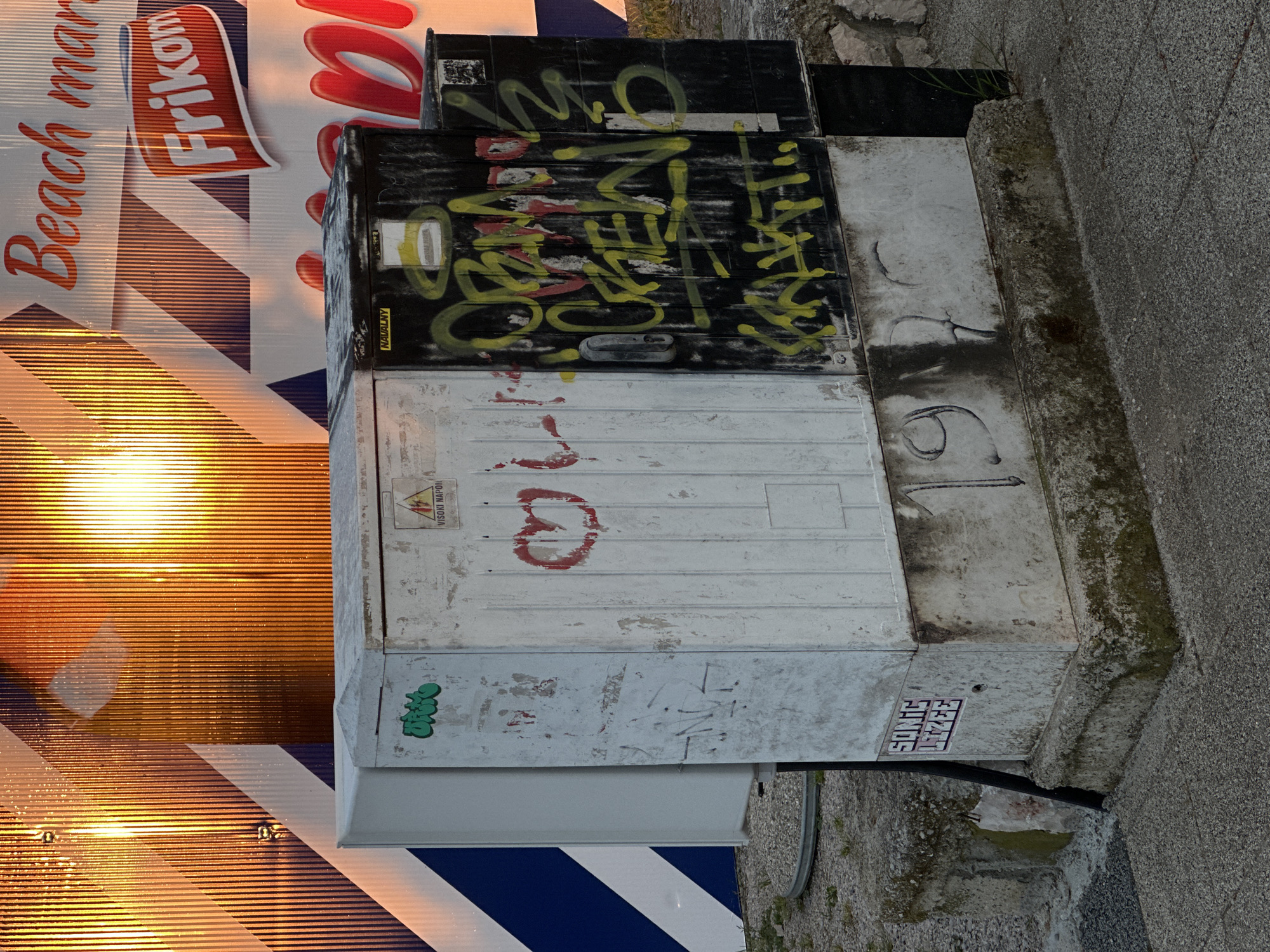 The image displays an outdoor scene in Šušanj, Montenegro, featuring commercial signage and heavily graffitied utility boxes. On the left, a large, corrugated advertisement or sign shows the partial text "Beach mar" (likely "Beach market") and the distinctive logo for "Frikon," a regional ice cream brand. A bright, warm light source, possibly an artificial light or the sun during golden hour, creates intense reflections and horizontal golden streaks across the lower section of this sign.
To the right, a stack of three weathered electrical or utility boxes is visible. The uppermost box is black and extensively covered in yellow and red spray paint, featuring various abstract tags and markings that are difficult to decipher. A small yellow sticker on this box reads "NAVALNY." The middle box, white in color, shows faded red graffiti, including a shape resembling a heart or the number "3." A sticker on this box contains the text "VISOKI NAPON" (High Voltage) alongside a lightning bolt symbol. The bottom box, also white, features green, bubbly graffiti that could be "GRAFF" or a similar stylized word, and another sticker with fragmented text like "SOKOLI" and "EZAJ." Faint, stylized numbers resembling "19" or "61" are also visible on one of the lower white panels.
The boxes appear dirty and worn, with traces of moss and grime on the adjacent concrete ground and wall to the far right. The scene is static, devoid of people or direct activity, presenting a typical urban environment blending commercial advertising with public infrastructure and street art.