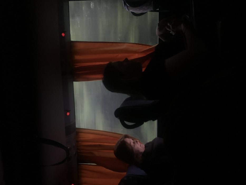 The image shows the interior of a bus. Two people are sitting in the seats, one facing the window and one facing the back of the bus. The person facing the window is a woman with dark hair, and the person facing the back of the bus is a woman with lighter hair. Both are sitting in the shadows, with only their outlines visible. The window is open and there are orange curtains on either side of the window. The window is blurry, suggesting it is raining or there is a lot of movement outside. The location of the image is in Łagów, Poland, based on the coordinates 51.382 latitude and 21.738 longitude. The image is dark, with only the outlines of the figures and the window visible. The scene is quiet and contemplative, with a sense of movement outside the bus and stillness inside. The lighting is dim, with only the faint light from the window and the overhead lights of the bus illuminating the interior. The image evokes a sense of isolation and introspection.