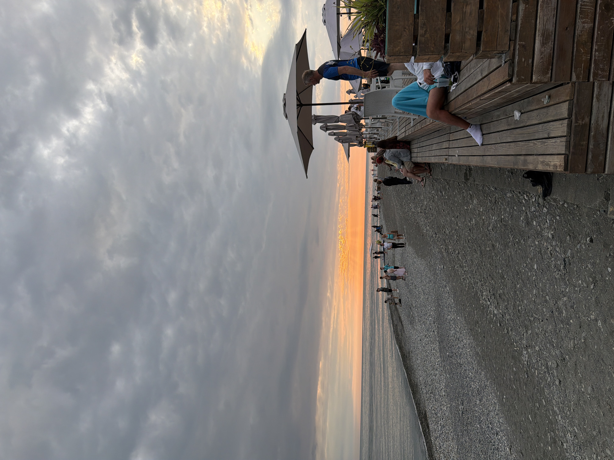 The image depicts a beach scene in Sirius, Russia, during a sunset. The sky is filled with dramatic, gray clouds, with a warm orange glow on the horizon, indicating the sun is setting. The beach is covered in small pebbles, and the water is calm.
On a wooden deck overlooking the beach, several people are seated and standing. One person is wearing a blue shirt and shorts, and another is wearing a white shirt and light blue shorts. There are several umbrellas and chairs set up on the deck, suggesting a cafe or restaurant.
Further down the beach, a group of people are walking and standing near the water's edge. The overall atmosphere seems relaxed, with people enjoying the evening by the sea. The time of day is clearly sunset, given the lighting and the warm colors in the sky. There is no visible text in the image.